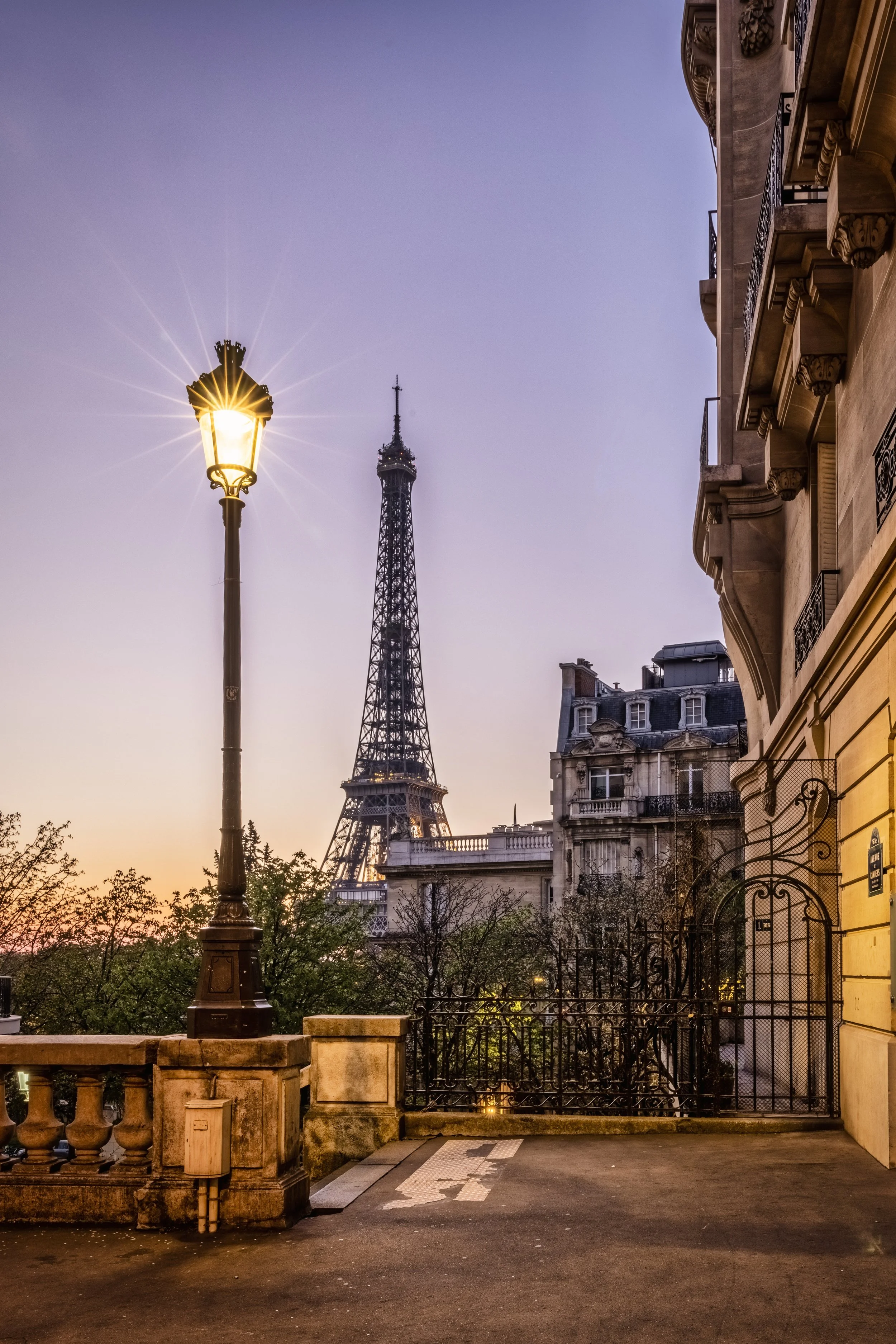 Eiffel Tower seen from a corner in Paris during sunset, with a street lamp in the foreground and buildings with ornate balconies on the right.