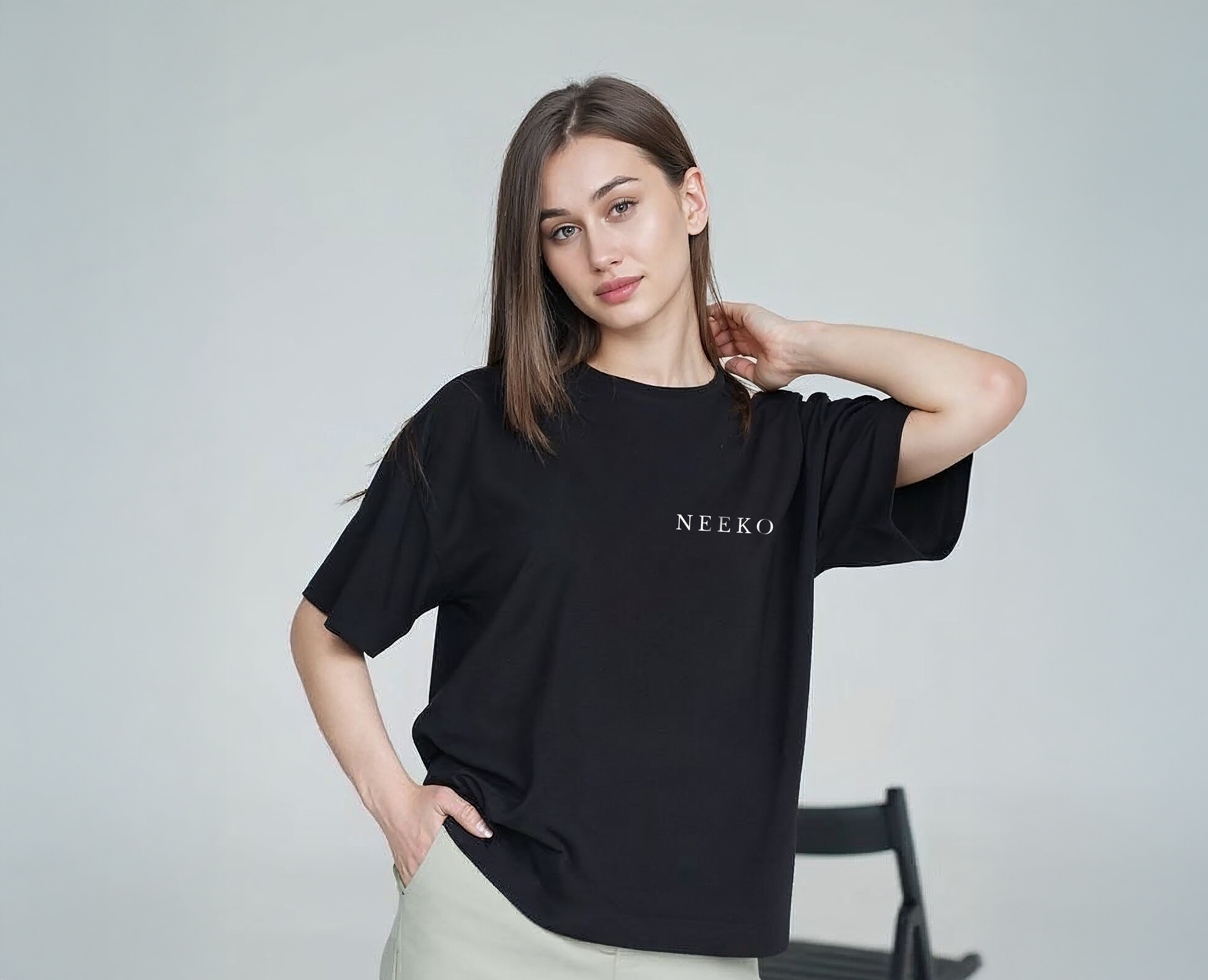 A young woman with straight brown hair, wearing a black T-shirt with the word NEEKO on it, poses with her right hand on her hip and her left hand touching the back of her head, standing against a light gray background.