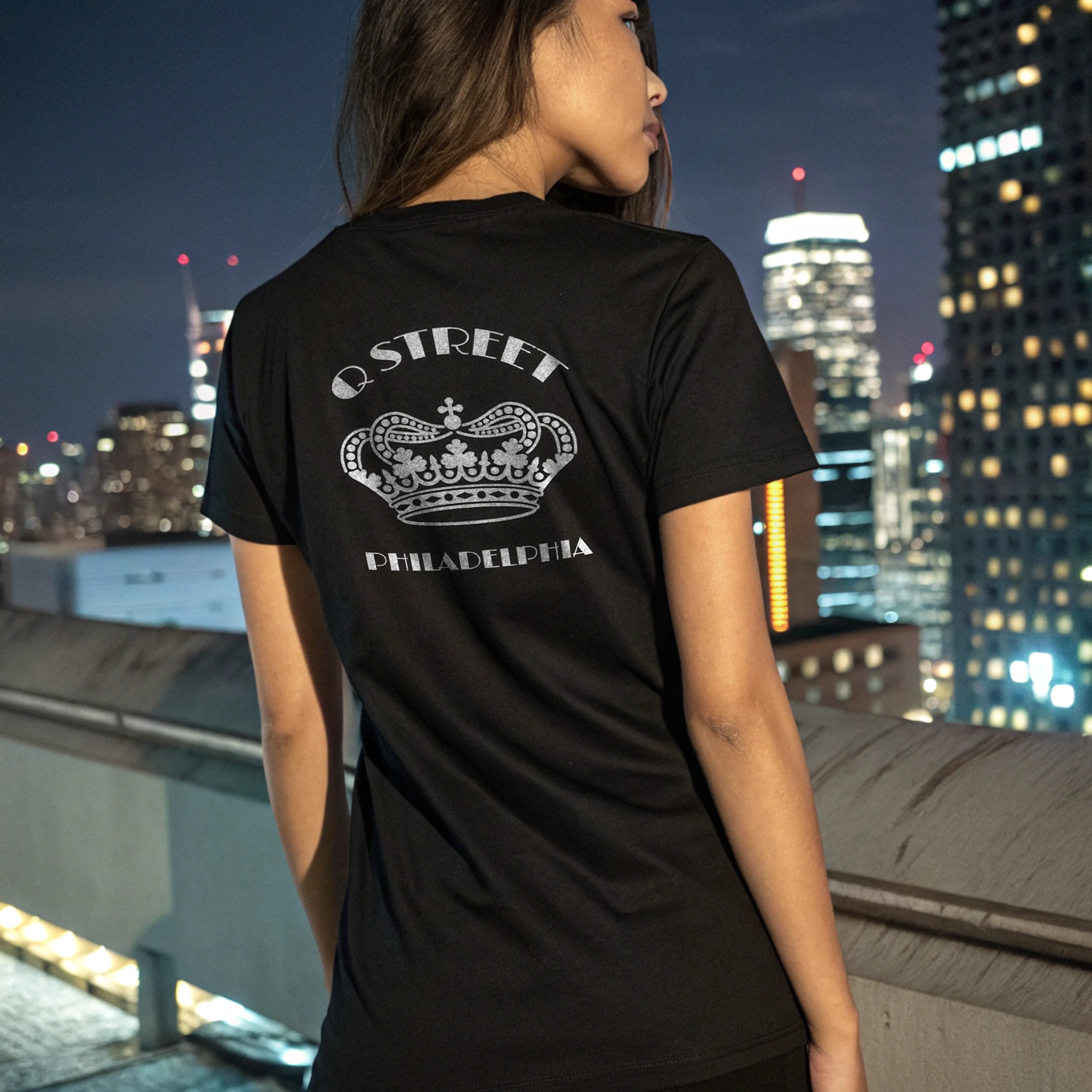 A woman is standing on a rooftop at night with a city skyline behind her. She is wearing a black T-shirt with a white royal crown graphic and the words 'Q STREET PHILADELPHIA' printed on the back.