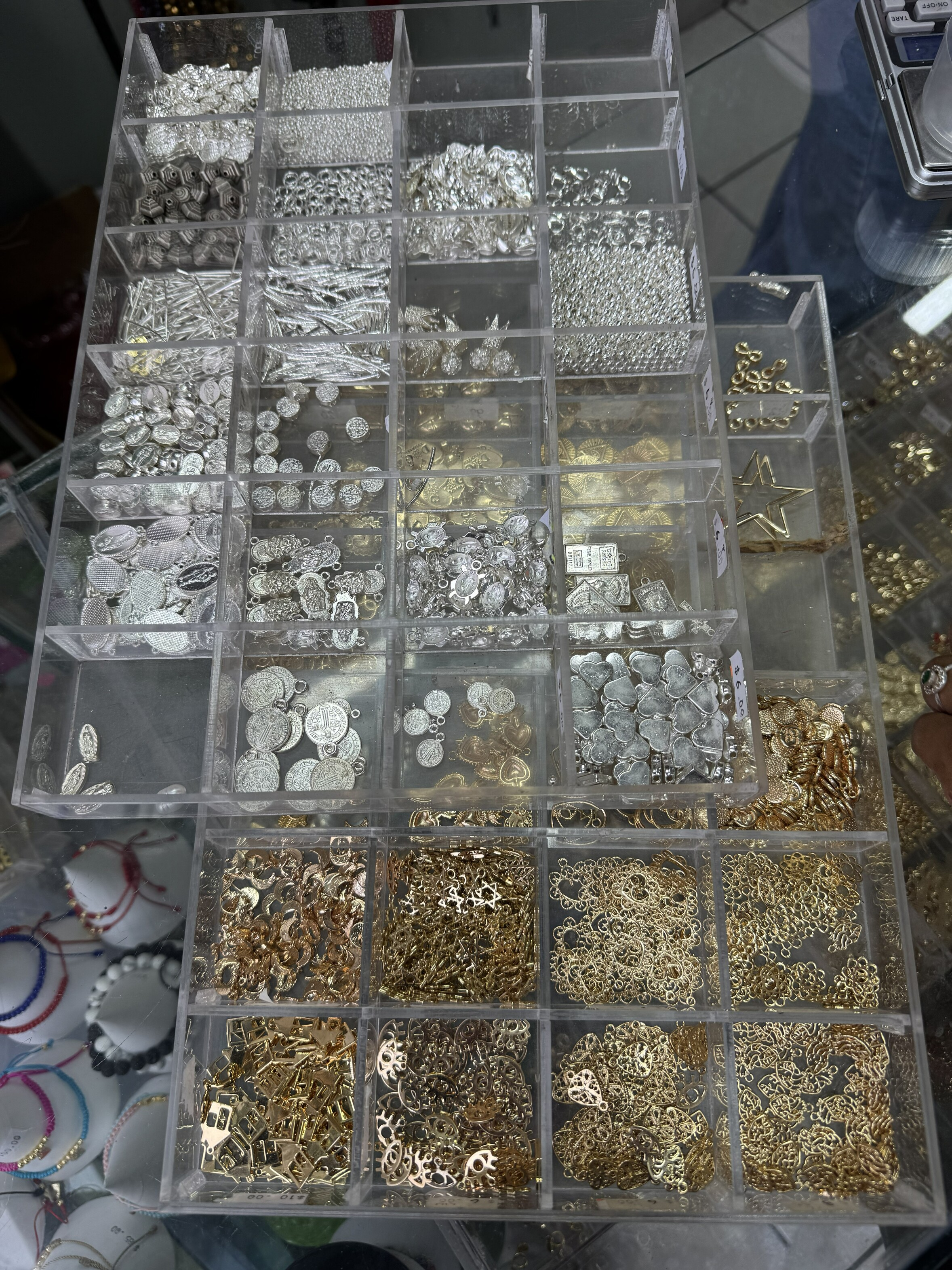 A display case with multiple compartments containing an assortment of shiny metallic jewelry charms and accessories in gold and silver colors.