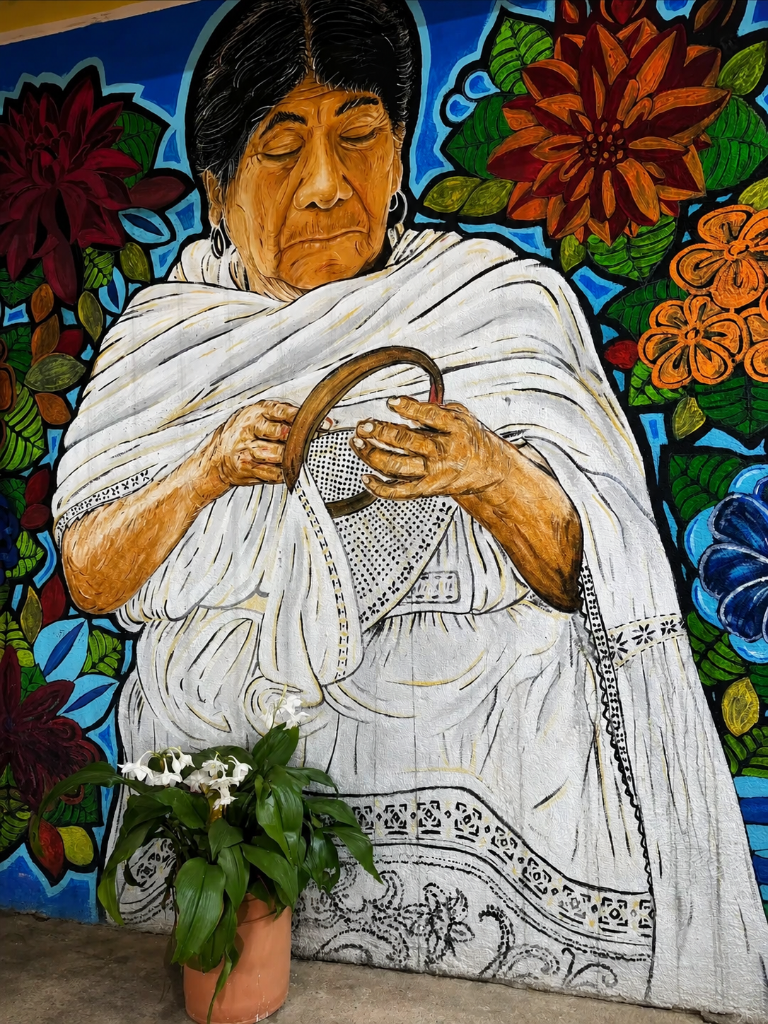A mural of an elderly woman with dark hair and earrings, dressed in a white garment, holding a small speaker, surrounded by colorful flowers. A potted white orchid plant is placed in front of the mural.