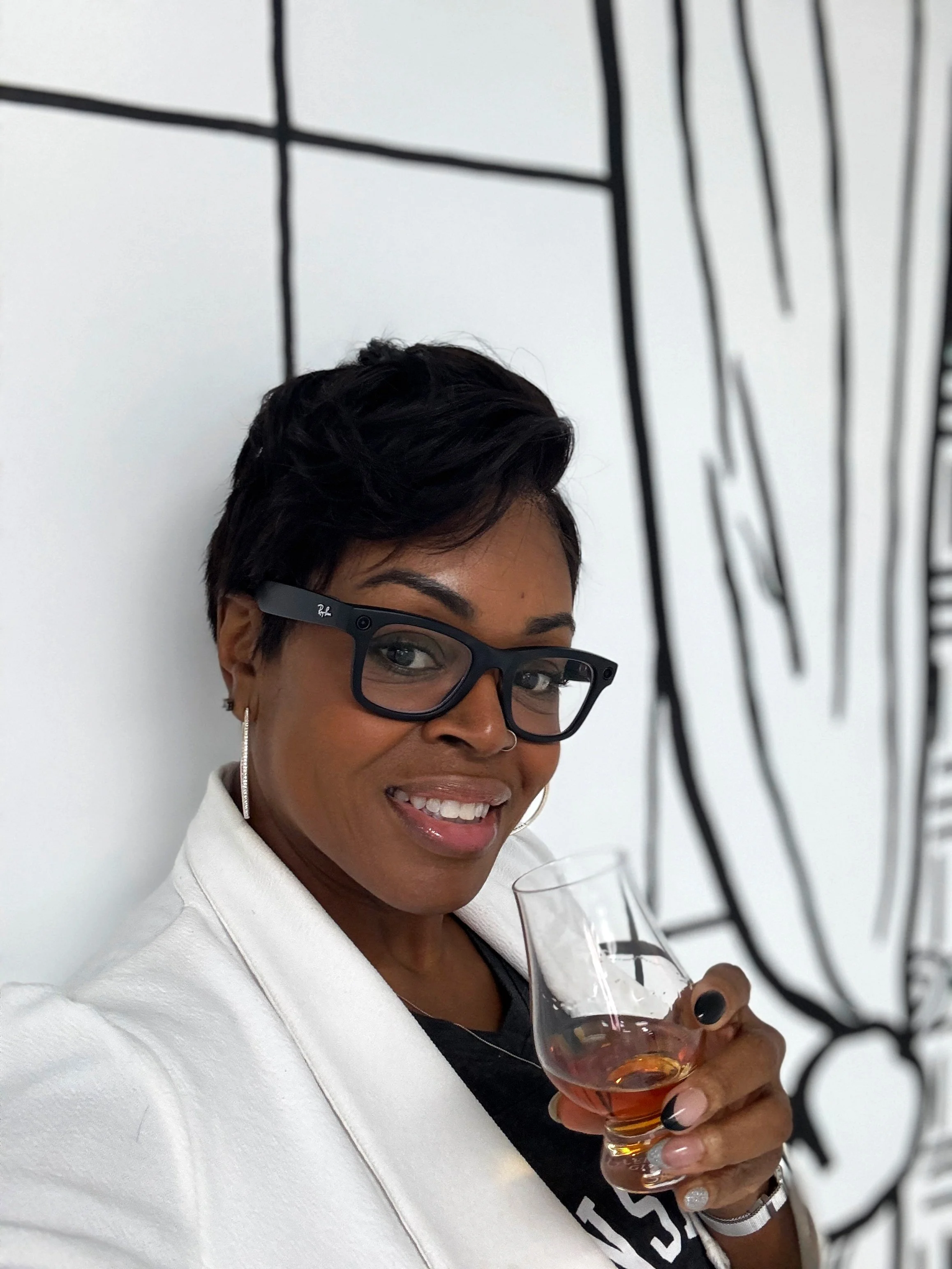 A woman with short black hair, glasses, and hoop earrings smiling and holding a glass of rosé wine, standing in front of a white wall with black abstract line art.