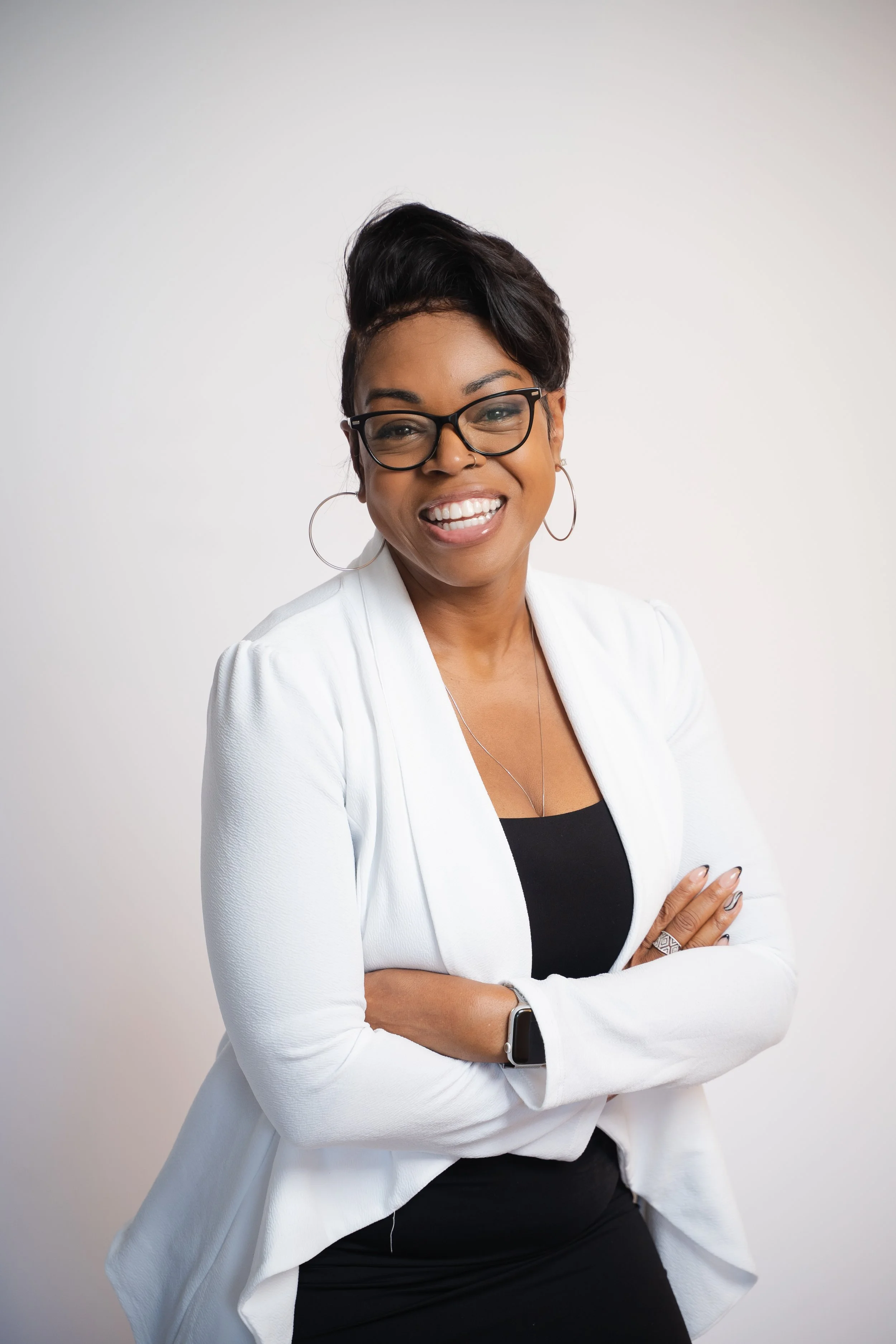 A smiling woman with glasses and hoop earrings, wearing a white blazer over a black top, standing against a plain white background.