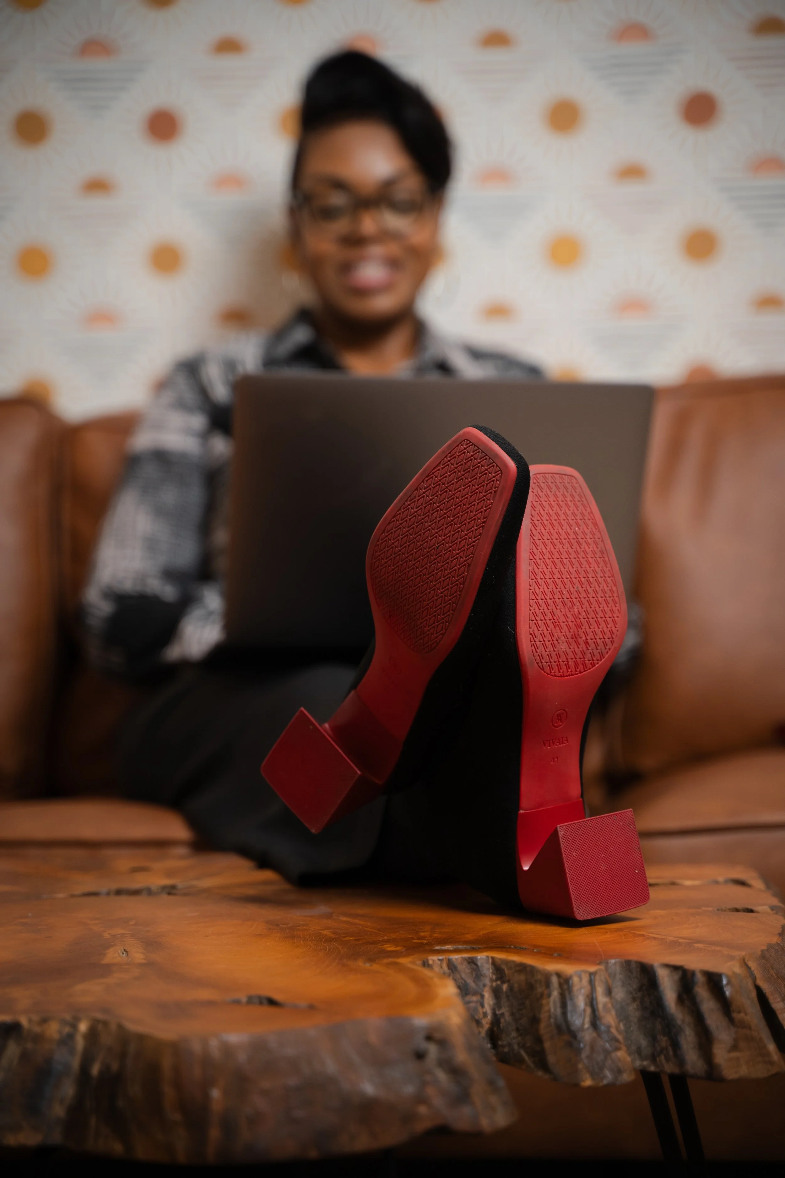 A woman sitting on a leather couch working on a laptop with red high heels prominently displayed on a wooden table in the foreground.