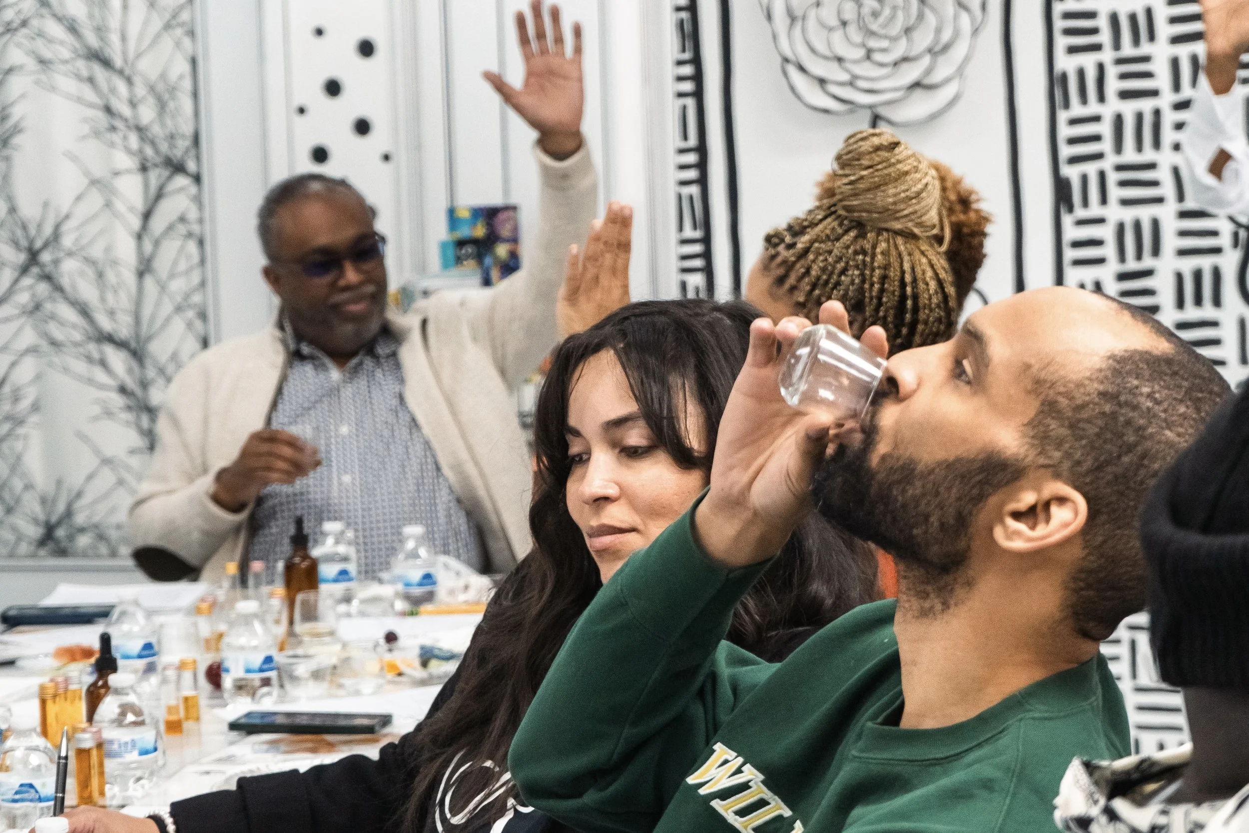 A group of people at a table, with one man drinking from a shot glass, and a woman looking down with a thoughtful expression. In the background, a man with gray hair and glasses raising his hand, surrounded by various bottles and items on the table.