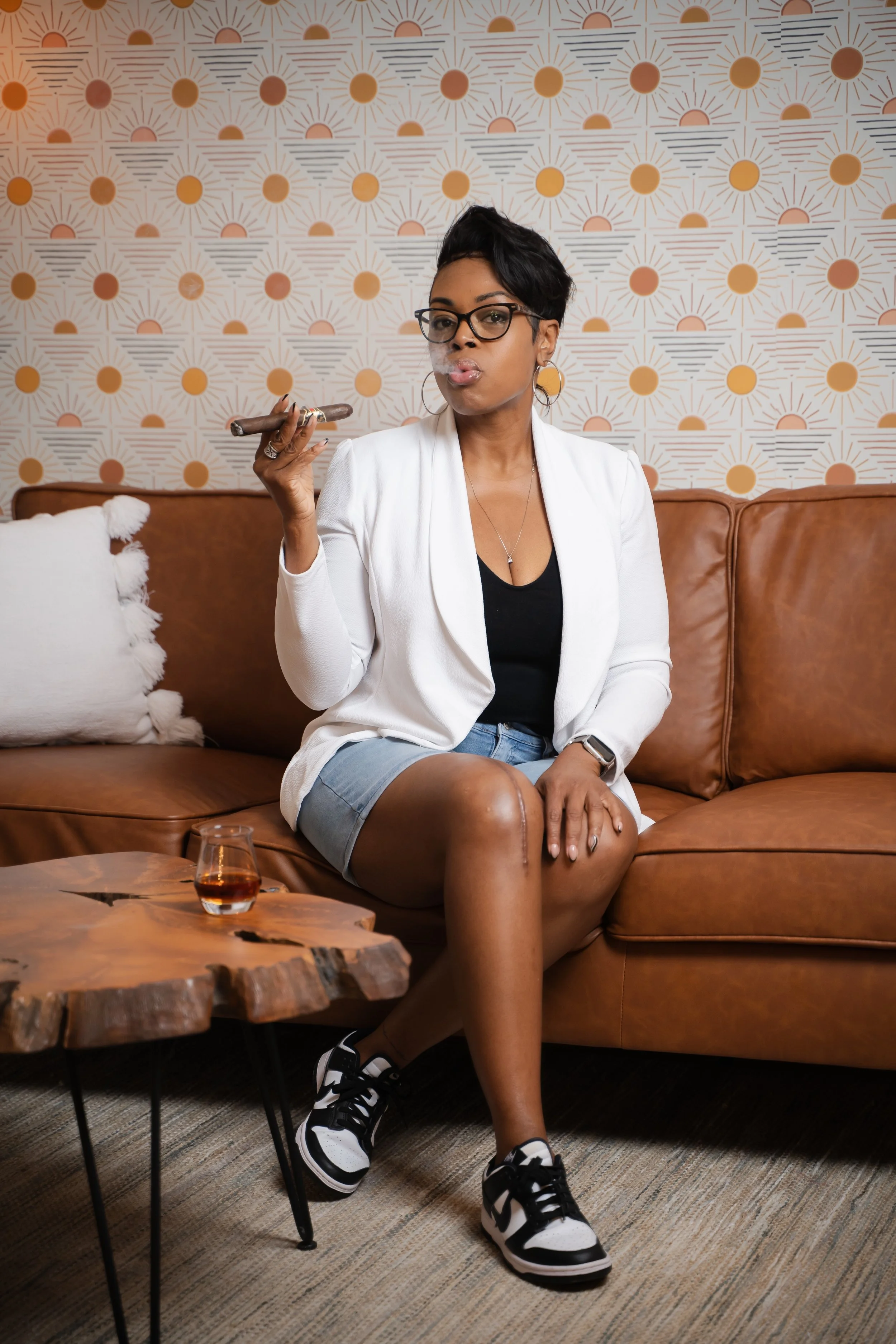 A woman with short black hair, glasses, and hoop earrings sitting on a brown leather sofa, smoking a cigar, dressed in a white blazer, black top, denim shorts, in a modern living room with patterned wallpaper, a wooden coffee table with a glass of whiskey, and a decorative pillow.