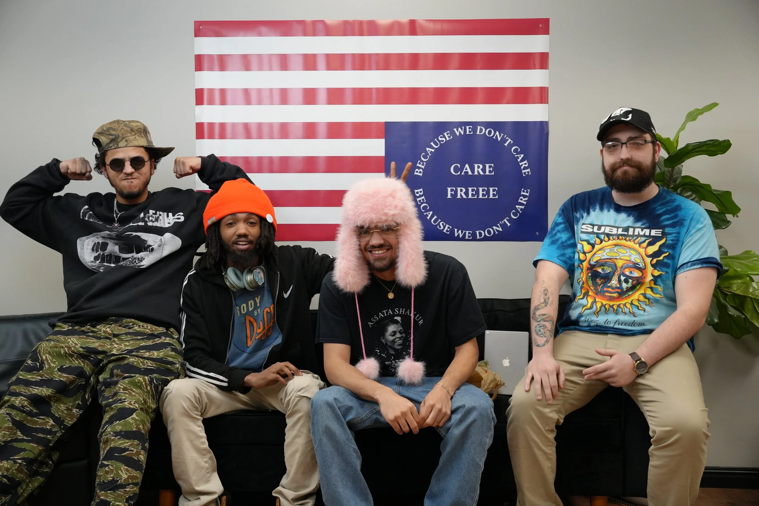 Four men sitting on a black couch in front of an American flag and a blue sign that reads 'Because we don't care, care free.' The man on the far left is wearing camouflage pants, a black sweatshirt, and a camouflage hat, flexing his arms. The second 