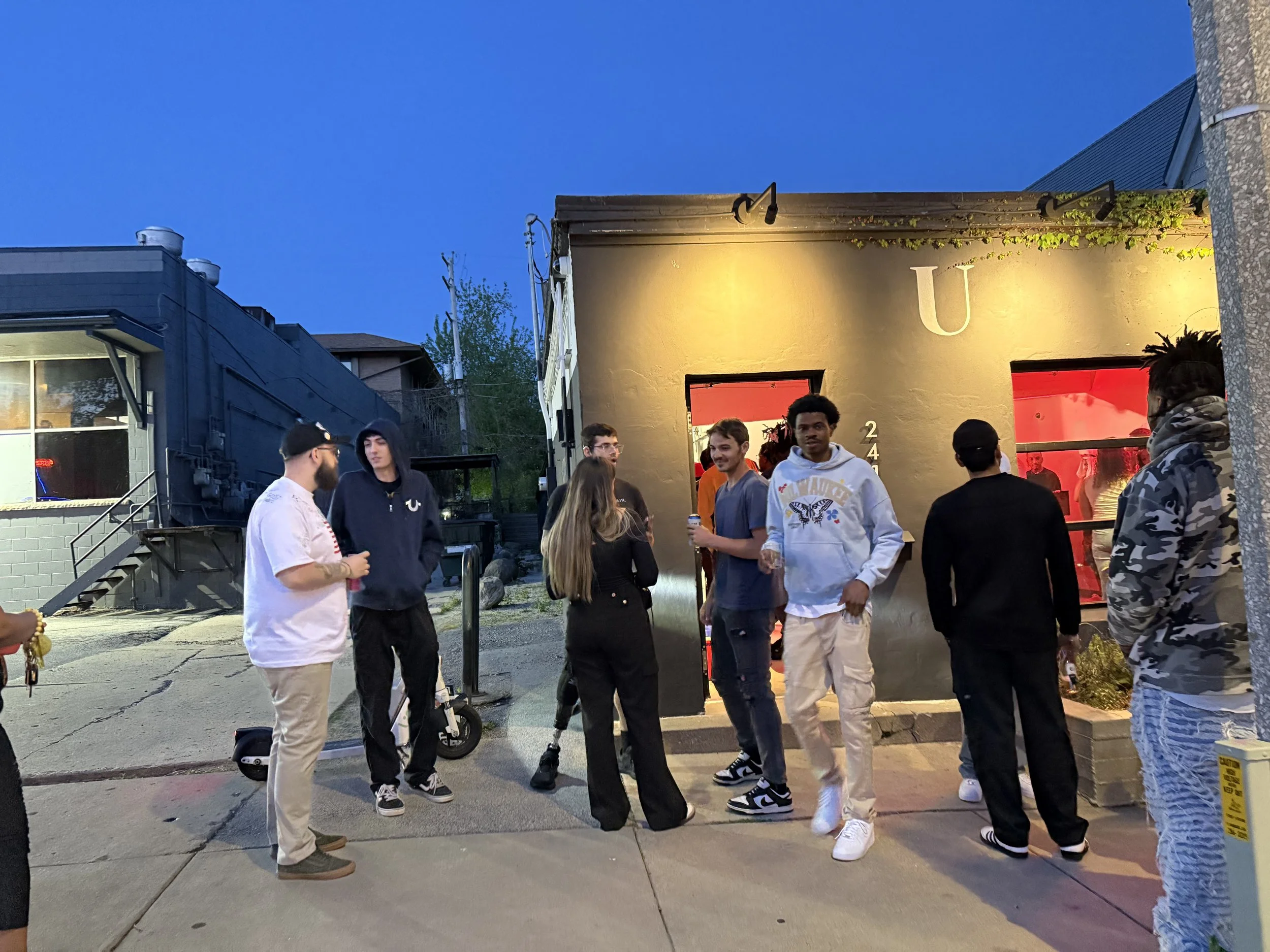 Group of people socializing outside a dark building with a large letter 'U' on the wall, during evening with a clear sky.