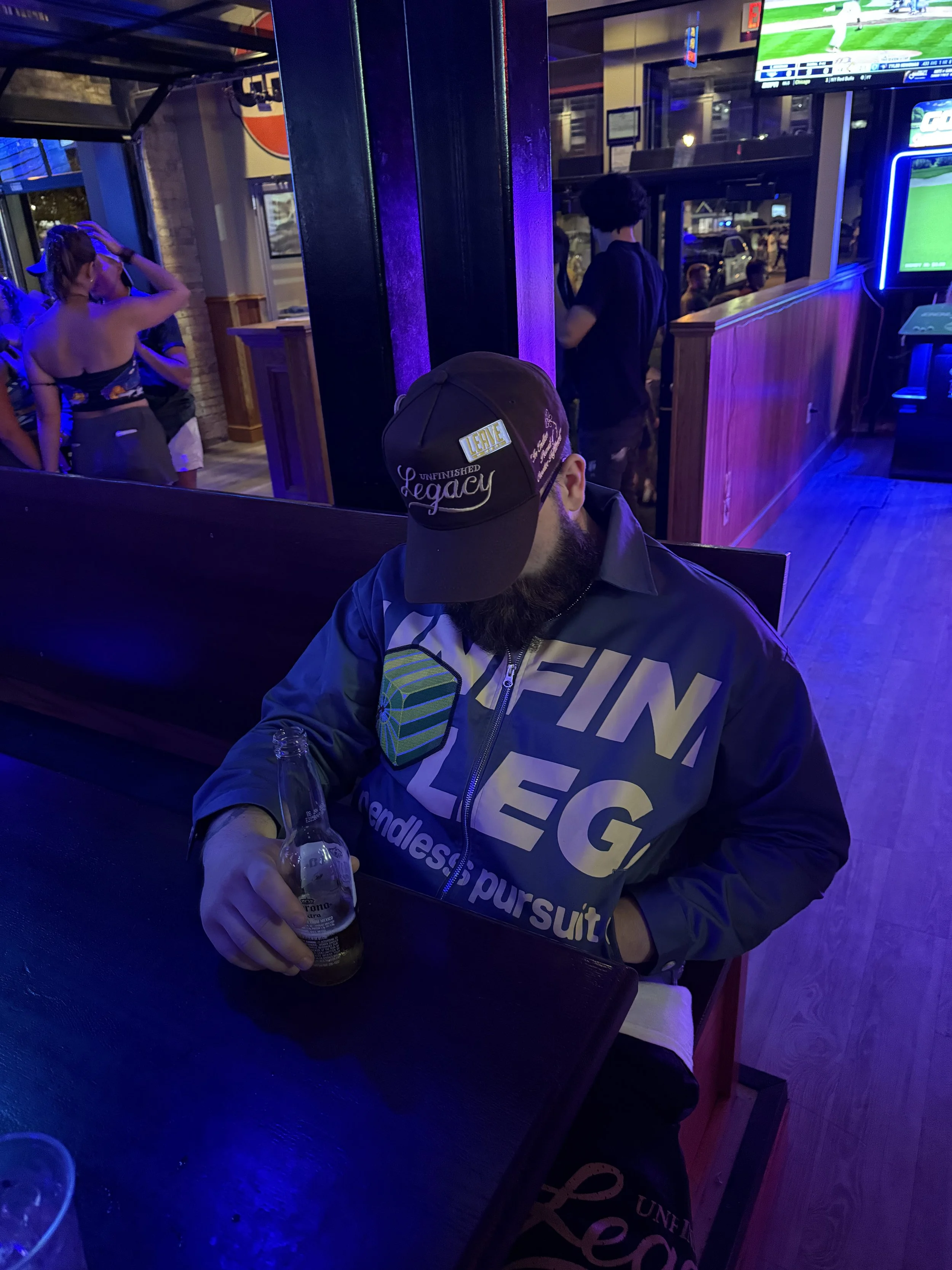 A man sitting at a bar table holding a beer bottle, wearing a dark jacket and a cap with text, in a bar with neon lighting and TV screens showing sports.