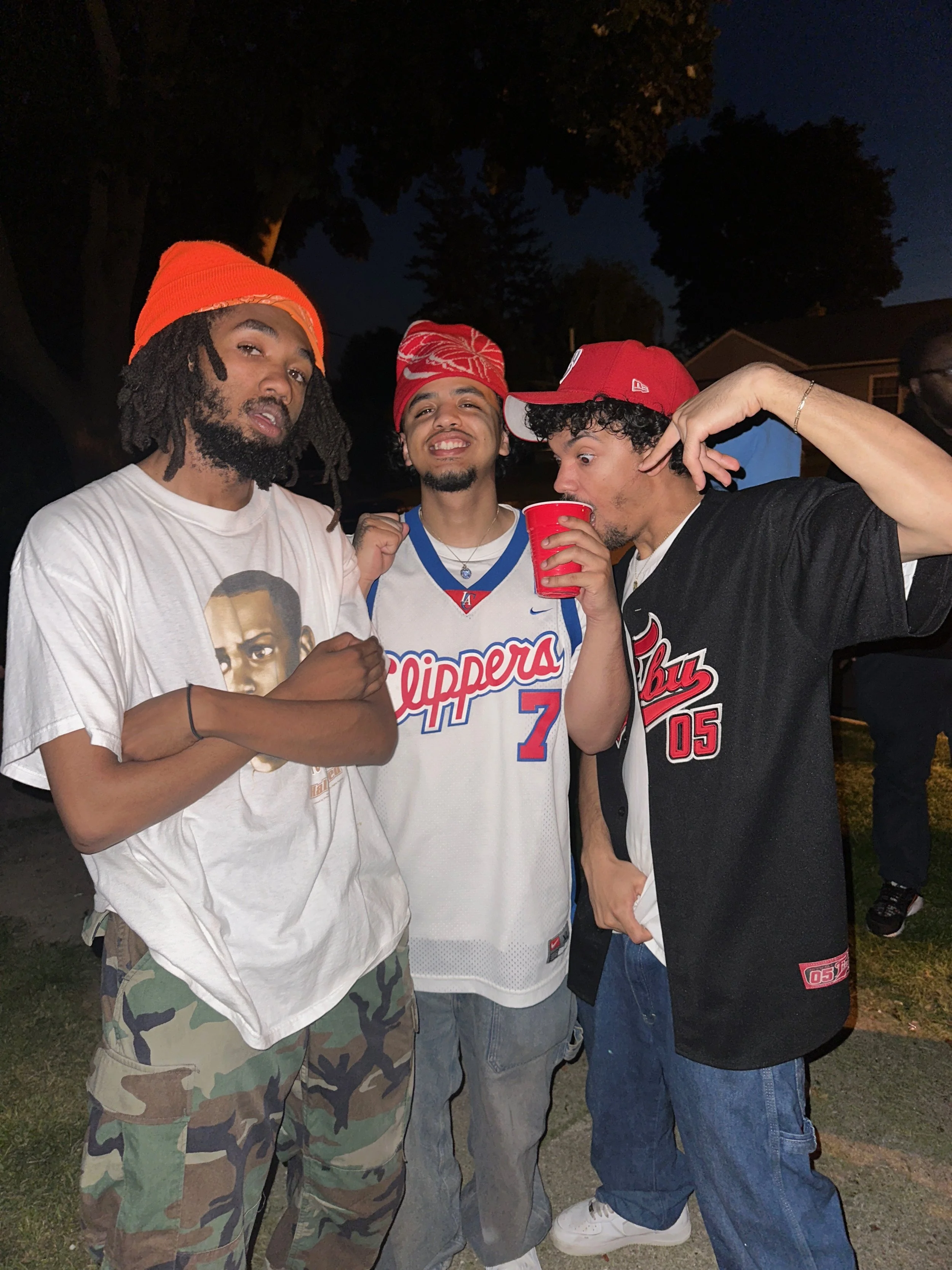 Three young men standing outside at night, smiling and posing for the camera; one is wearing a white t-shirt with a graphic, camouflage pants, and an orange beanie; the second is wearing a Los Angeles Clippers basketball jersey and a bandana, holding