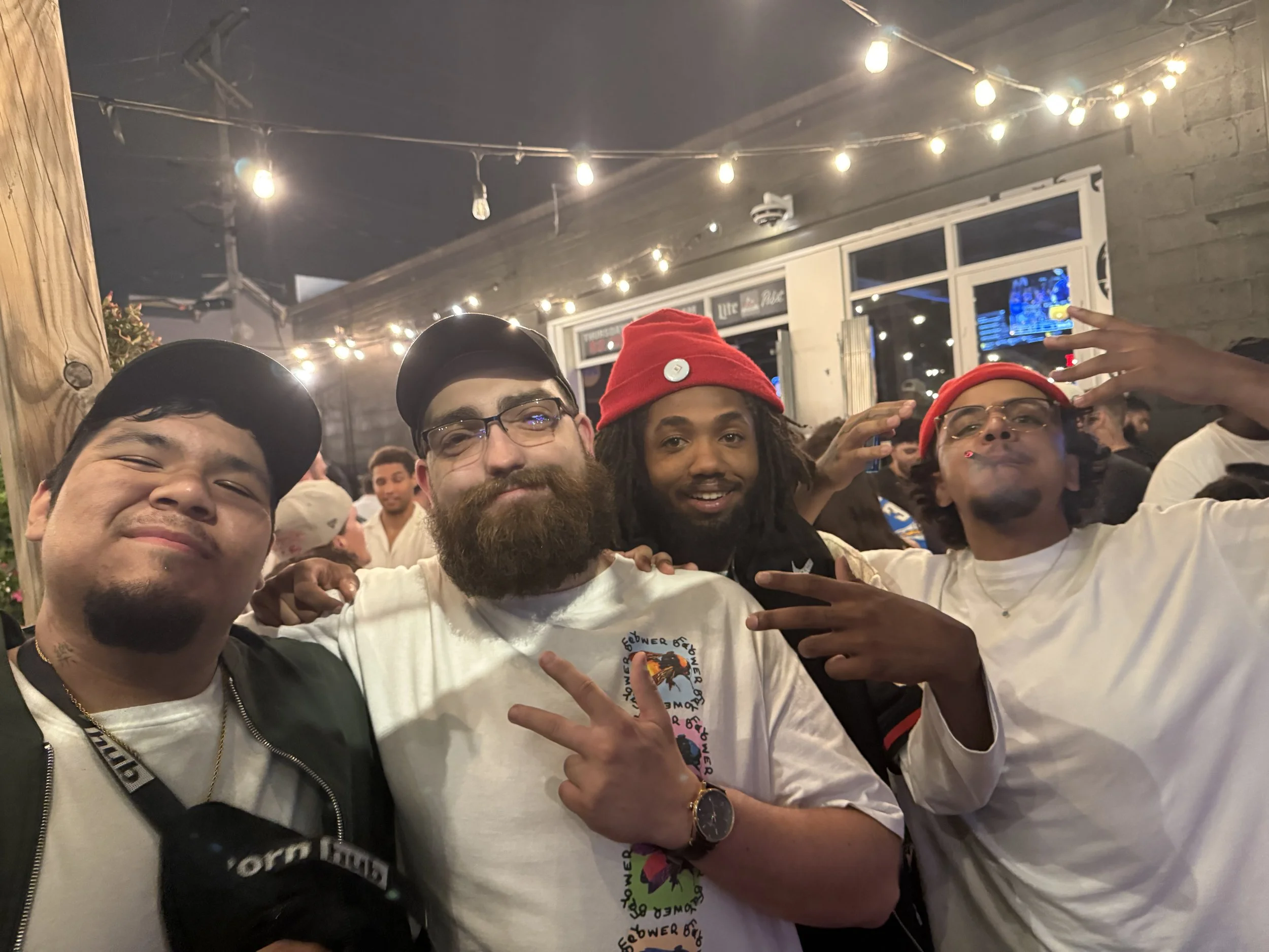 Four young men posing together at a party or gathering, smiling and making hand gestures, in an indoor space with string lights and people in the background.