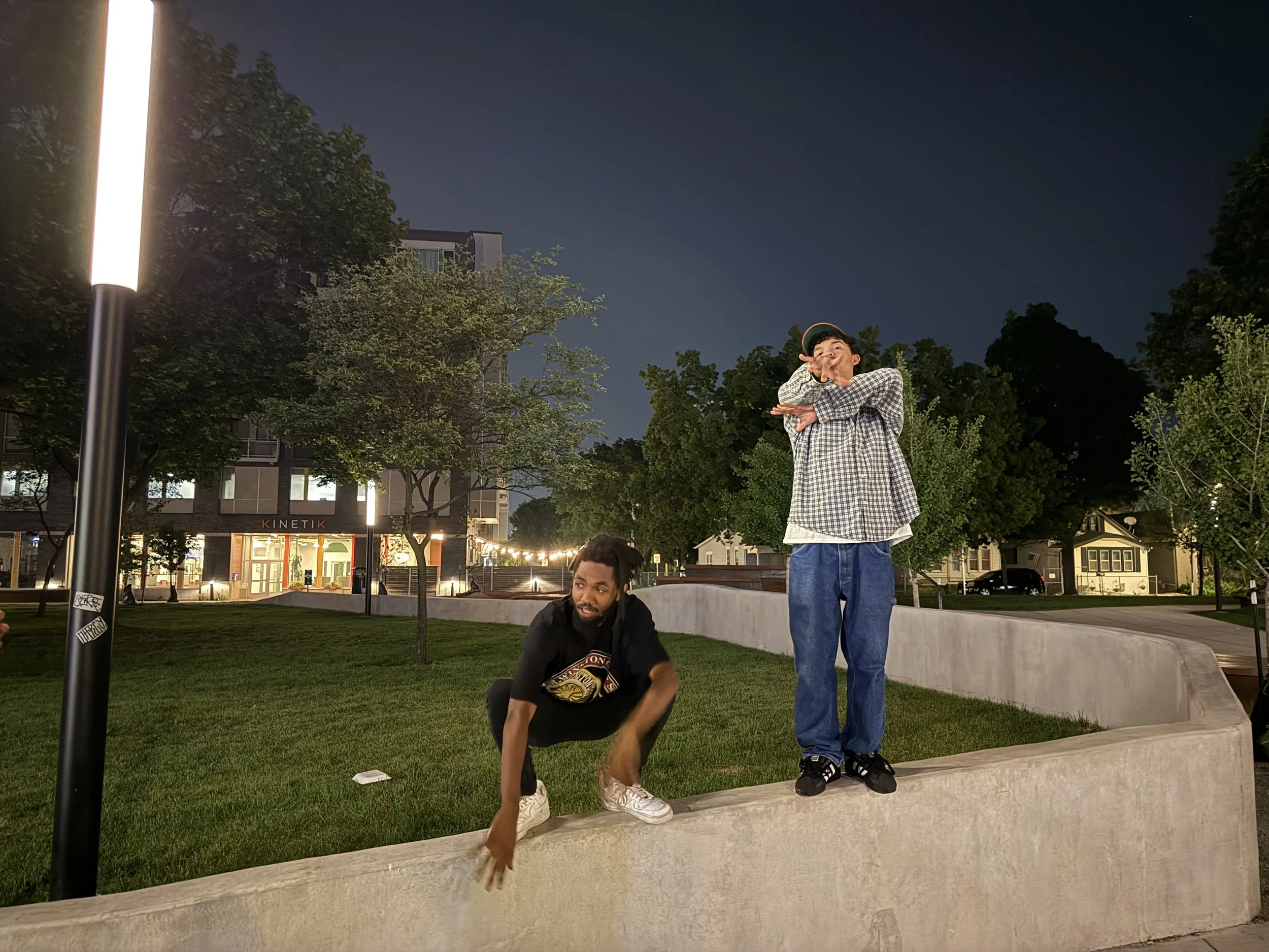 Two young men hanging out on a low concrete wall in a park at night, with trees and a building in the background. One is squatting and reaching down with his hand, the other is standing and pointing with his finger. The scene is illuminated by a stre