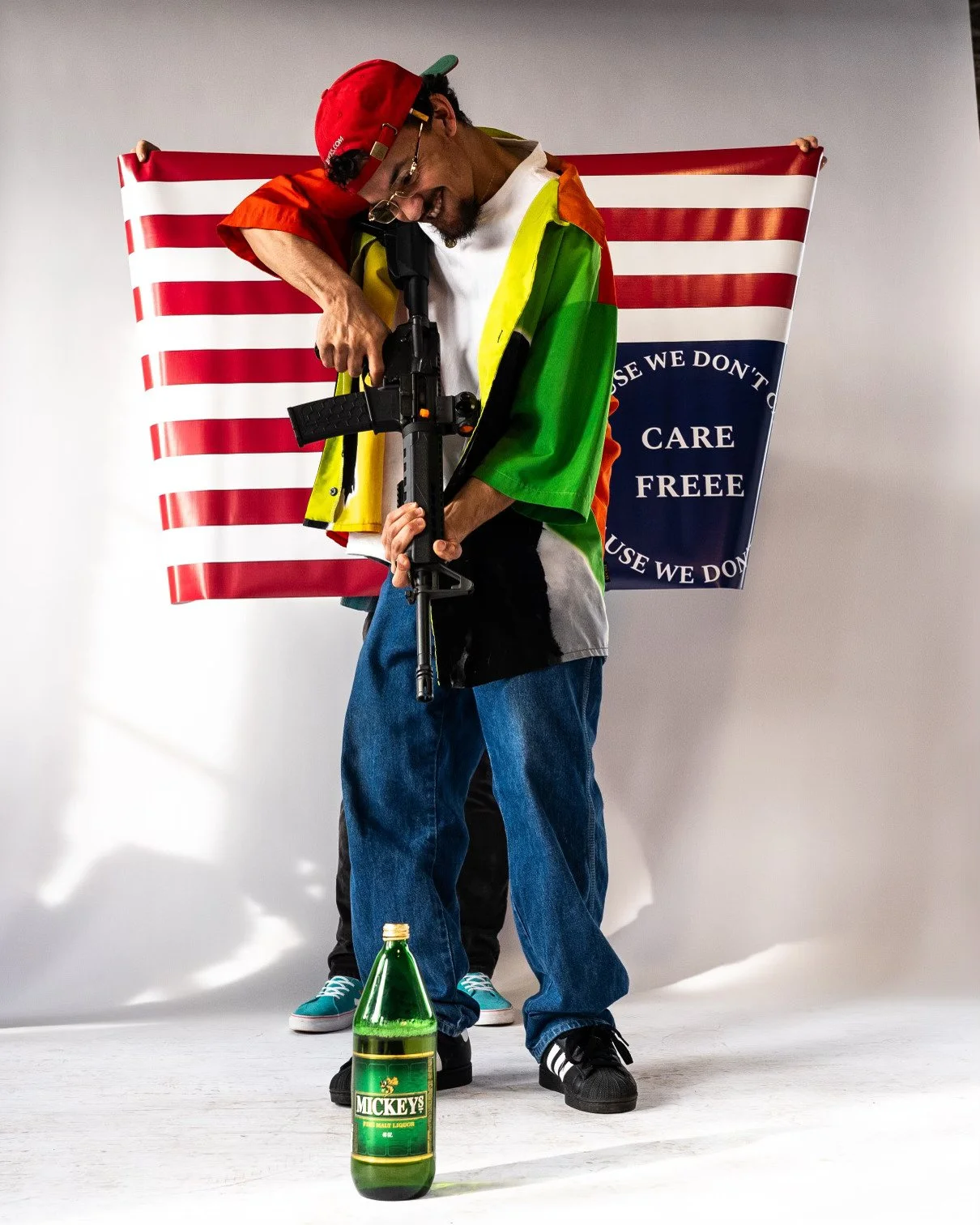 A man smiling and dressed casually in jeans, a colorful rainbow cape, and a red cap, holding a rifle in a studio with a large American flag in the background. There is a green bottle of Mickey's beer on the floor in front of him.