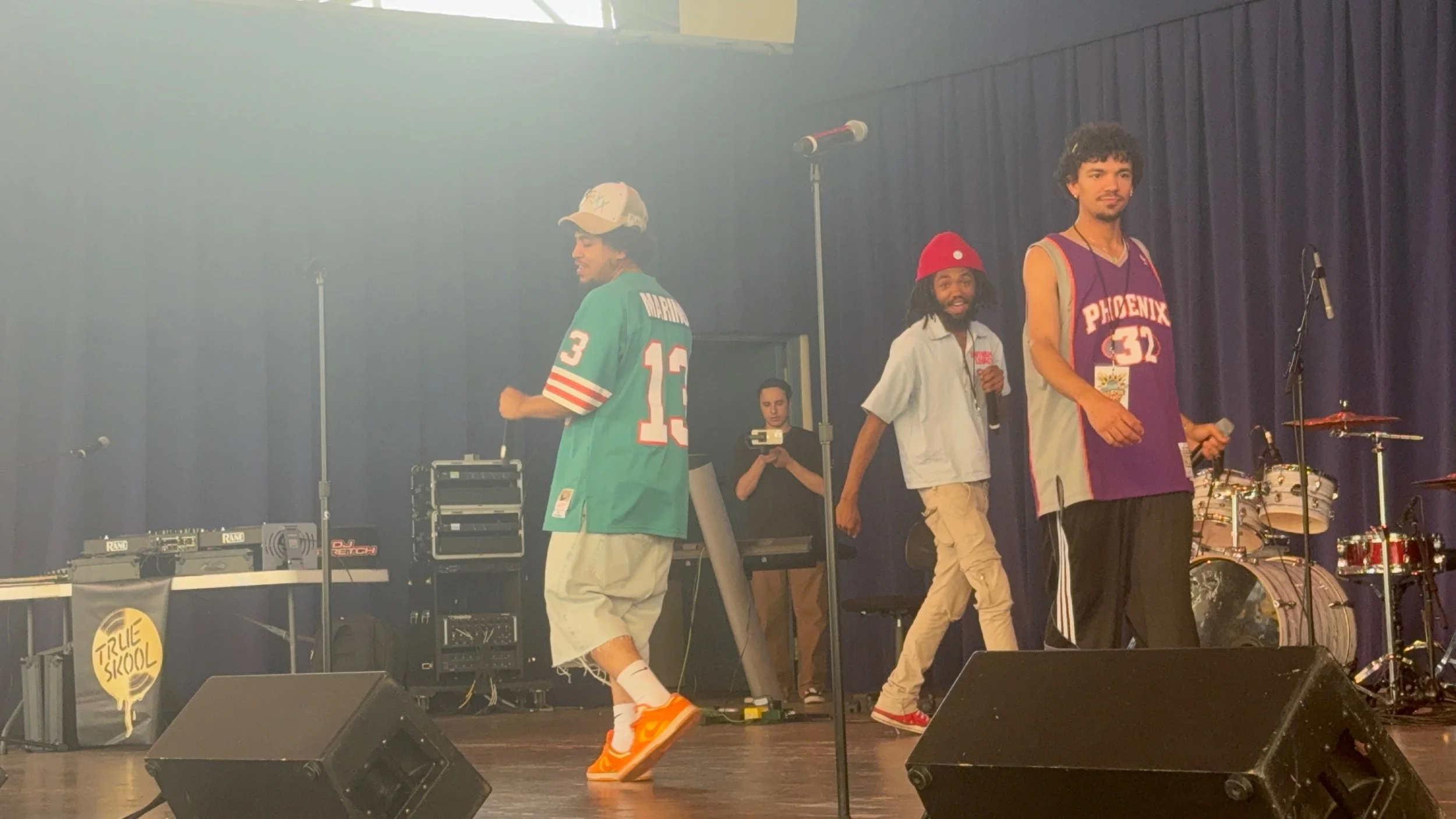 Three young male performers on stage, one wearing a teal jersey, the second in a gray jacket and red hat, the third in a purple Phoenix jersey, with musical instruments in the background and a person recording them.