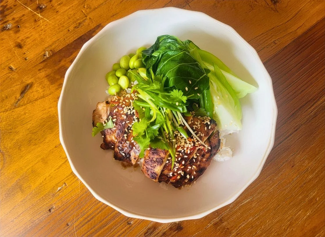 Grilled meat topped with sesame seeds and cilantro, served with fresh greens and peas in a white bowl on a wooden table.