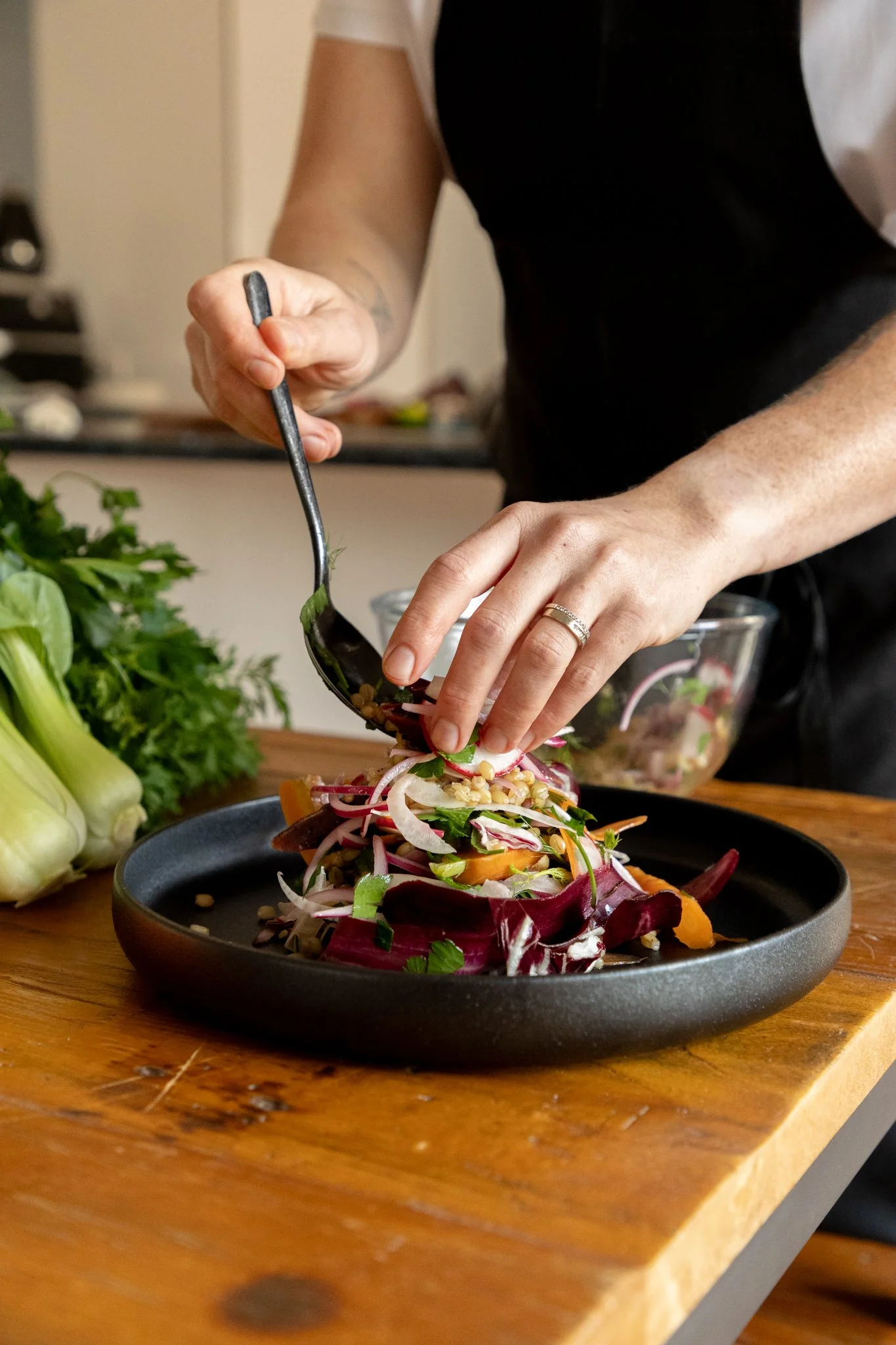 Person adding fresh salad ingredients to a plate on a wooden table, with herbs and vegetables in the background.