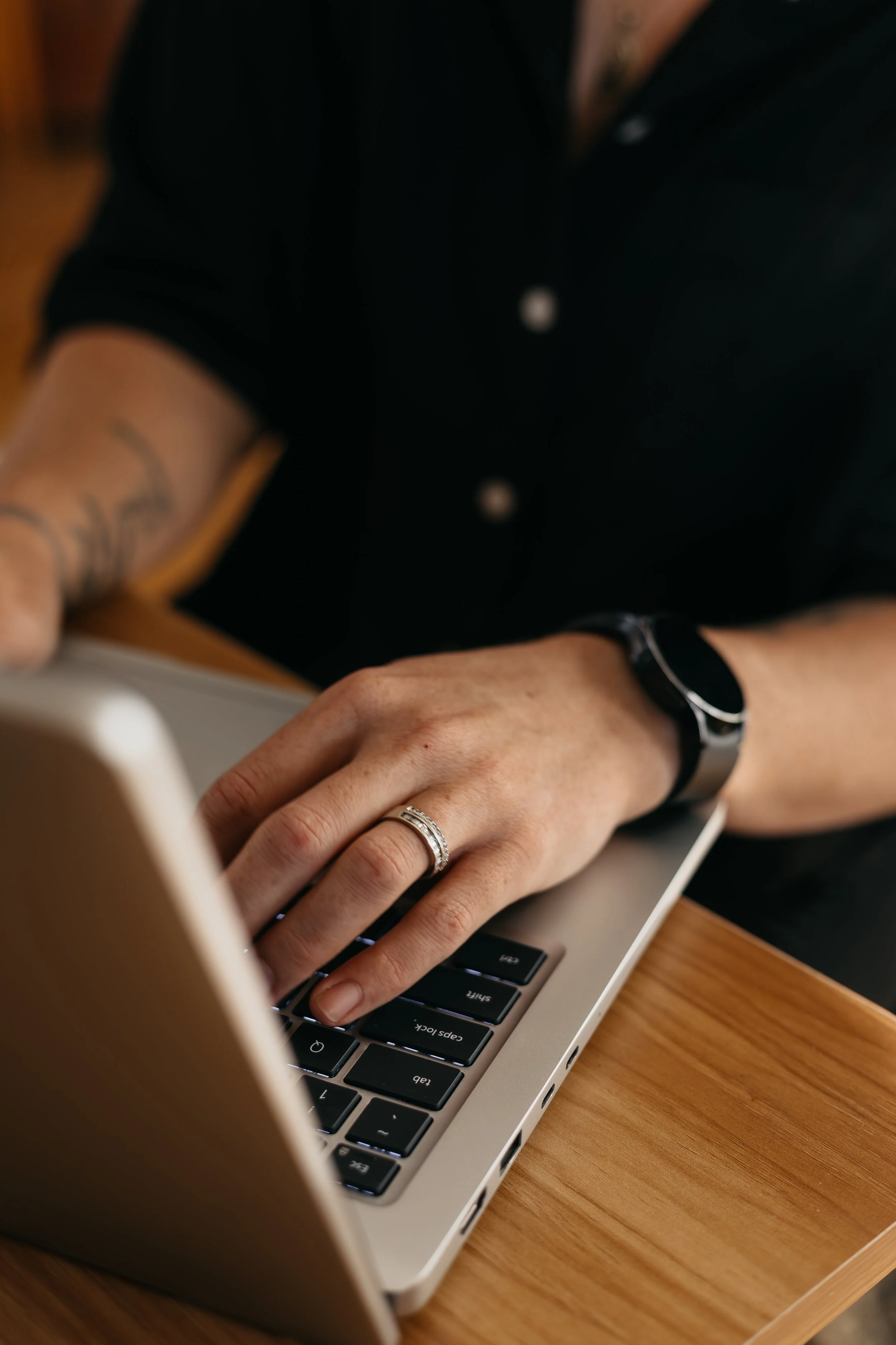 Close-up of a person's hands typing on a laptop keyboard at a wooden desk. The person is wearing a black watch and a ring, and has a tattoo on their arm. The background is blurred.