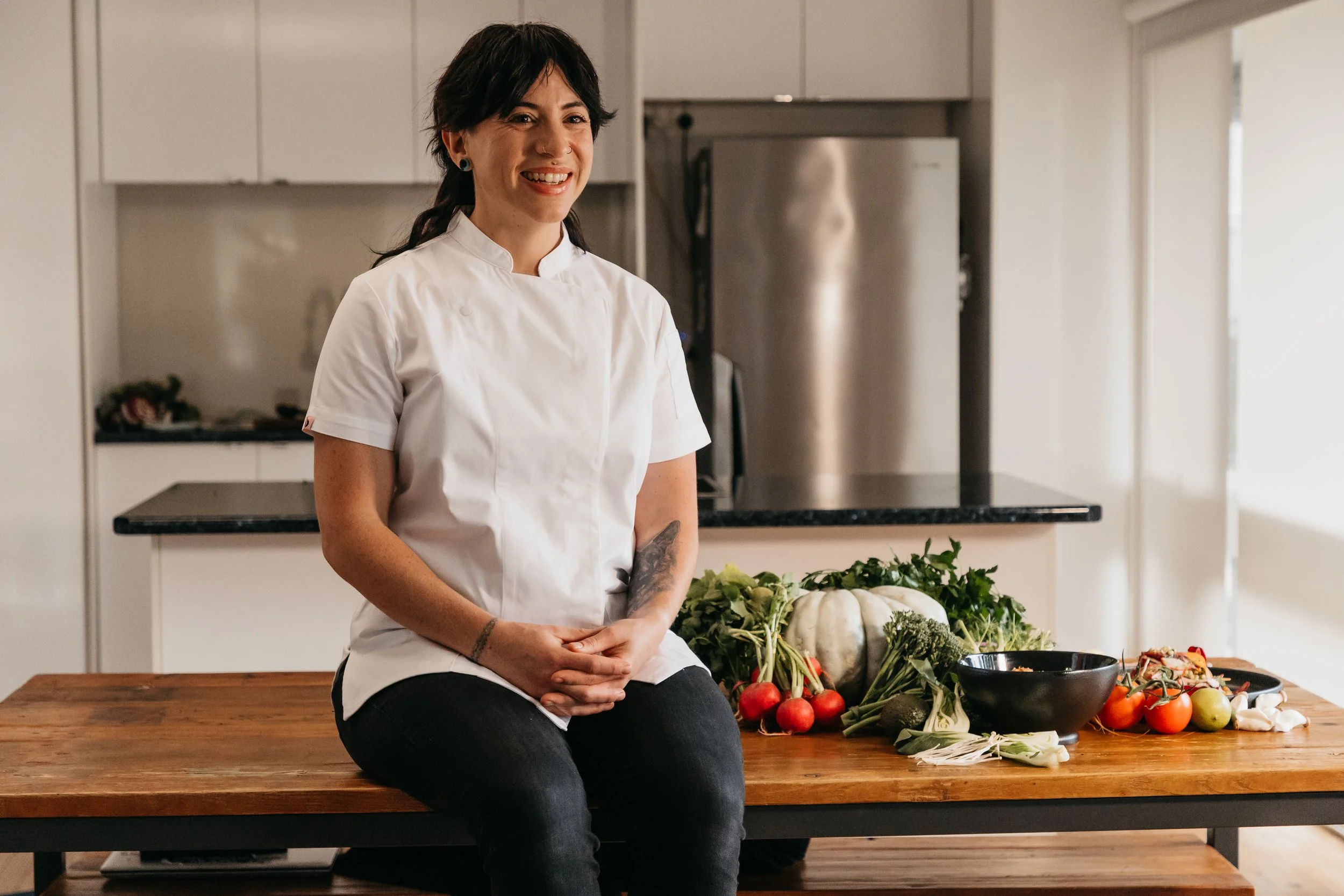 A woman in a chef's coat sitting on a wooden table with vegetables and herbs, smiling in a modern kitchen.