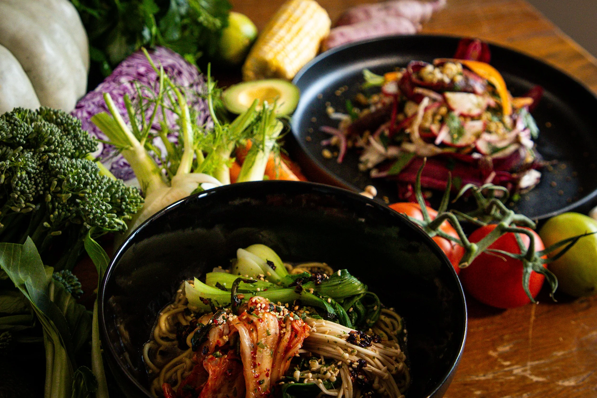 A bowl of Korean-style spicy noodles topped with sesame seeds and red pepper flakes, surrounded by fresh vegetables including broccoli, avocado, corn, tomatoes, and sweet potatoes on a wooden surface.