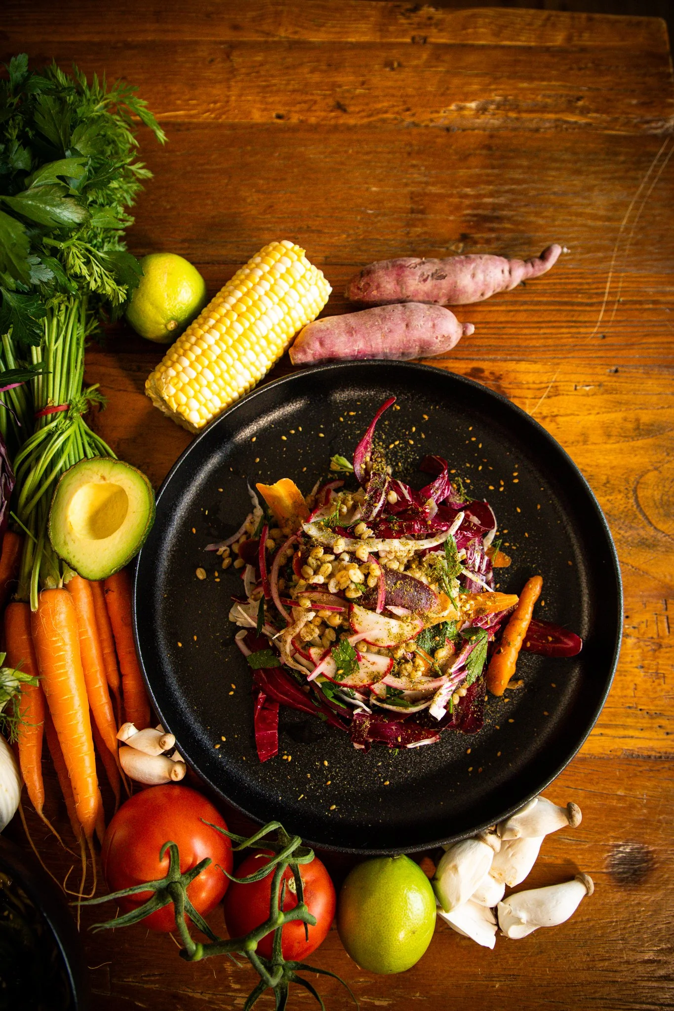 A black plate with a salad containing sliced radicchio, shredded carrots, chopped herbs, and sprinkled seeds. Surrounding the plate are fresh vegetables including carrots, tomatoes on the vine, green and yellow limes, an avocado, purple sweet potatoes, garlic, and mushrooms, all placed on a wooden surface.