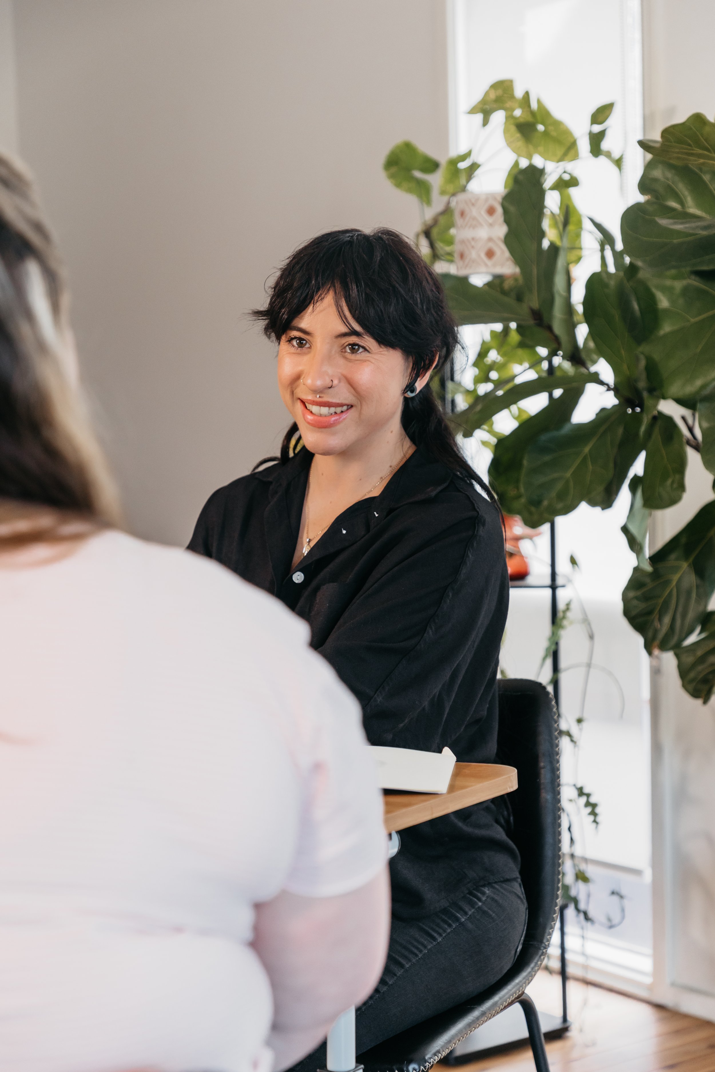 A woman with short black hair and a nose ring smiling and talking to another person in a bright room with large green plants and natural light.