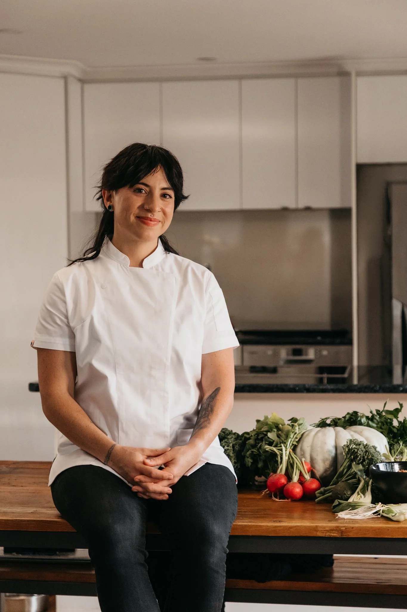 A woman wearing a white chef's coat sitting on a kitchen counter surrounded by vegetables including radishes, pumpkins, broccoli, and leafy greens.