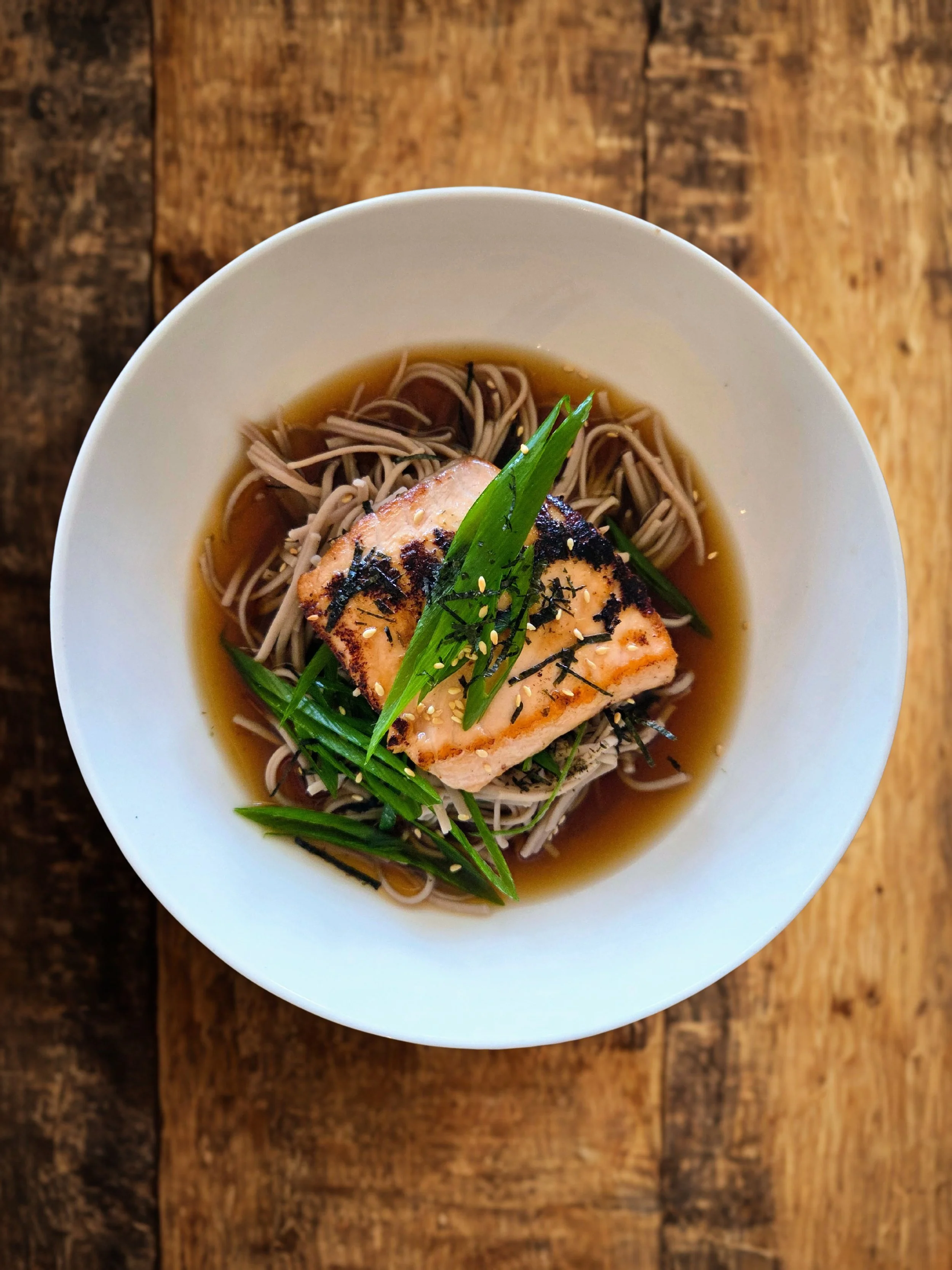 Bowl of ramen with grilled salmon, green onions, seaweed strips, and broth, on a wooden table.