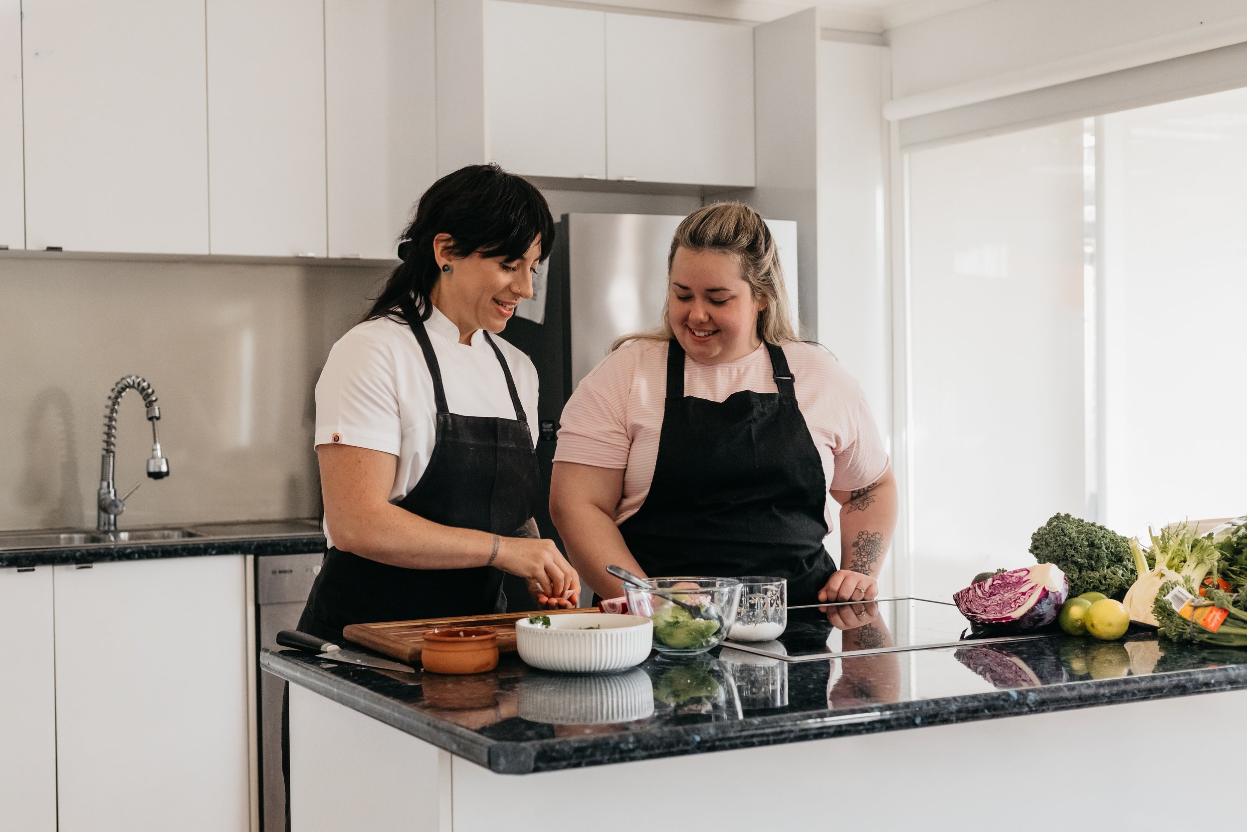 Two women with dark and blonde hair in black aprons preparing food together in a bright modern kitchen with white cabinets, a black countertop, and an assortment of fresh vegetables on the counter.