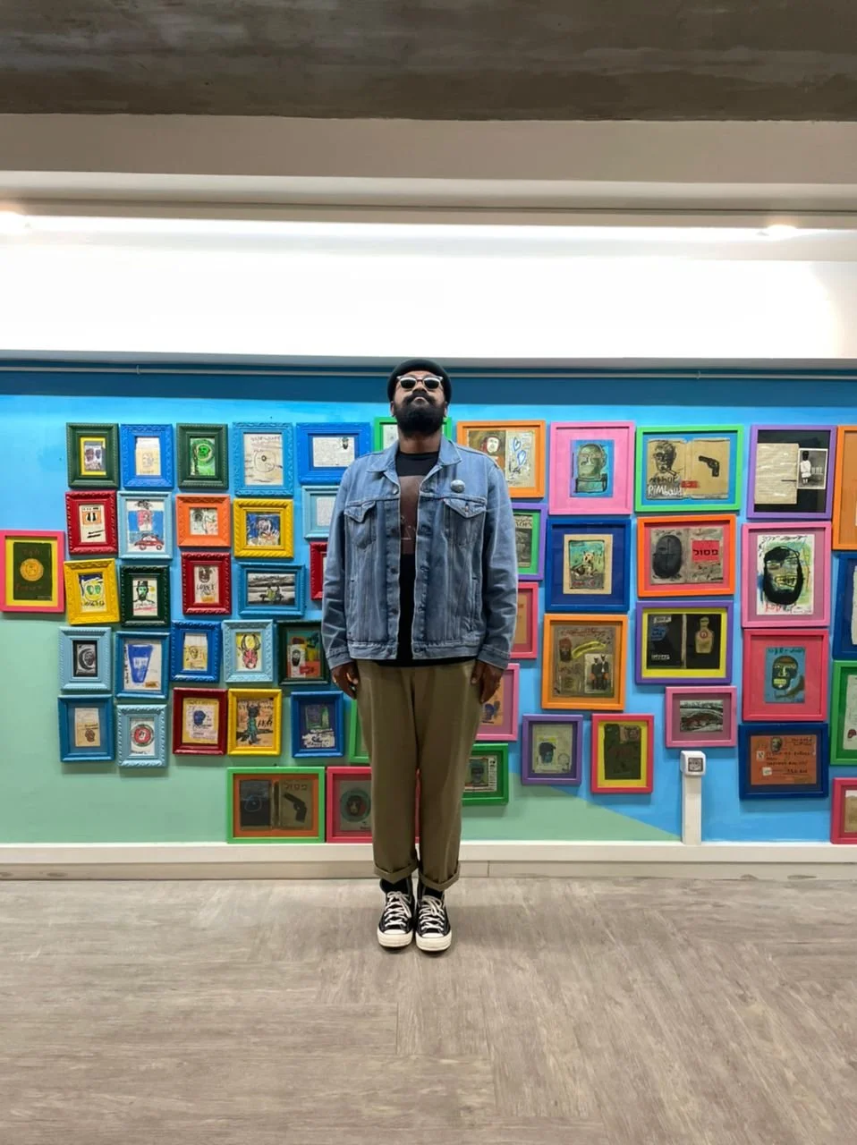 A man with a beard, wearing sunglasses, a denim jacket, a black shirt, beige pants, and black and white sneakers, standing in front of a colorful wall of framed artwork at an art gallery.