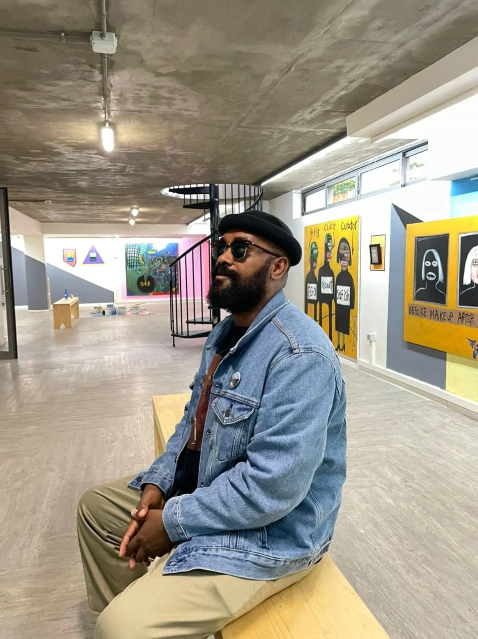 A man with a beard and sunglasses sitting on a wooden bench in an art gallery, wearing a black beret, denim jacket, black t-shirt, and beige pants, with colorful artworks on the walls behind him.