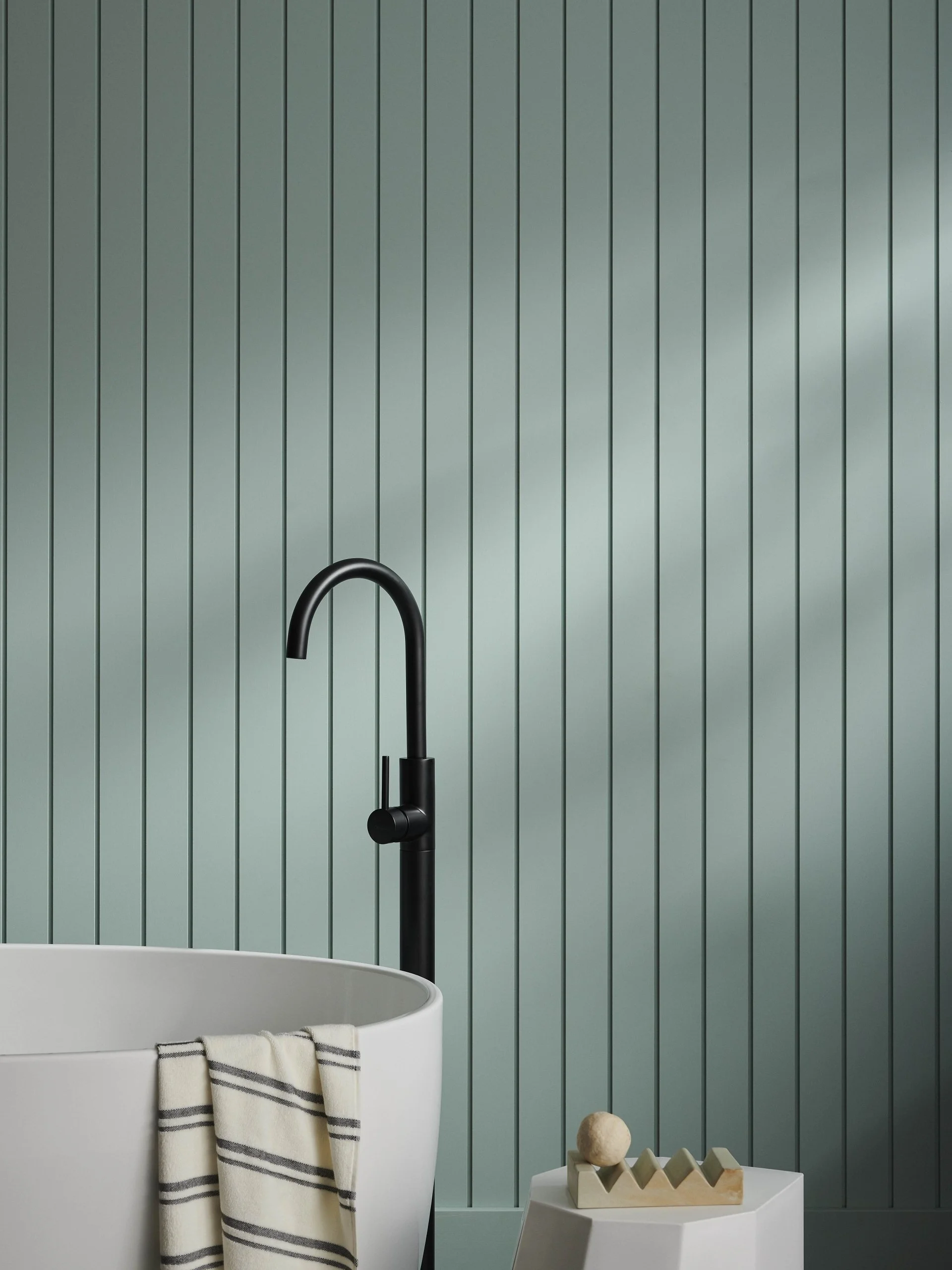 Bathroom with teal vertical paneled wall, black faucet, white bathtub with a striped towel, and small decorative objects on a white stool.