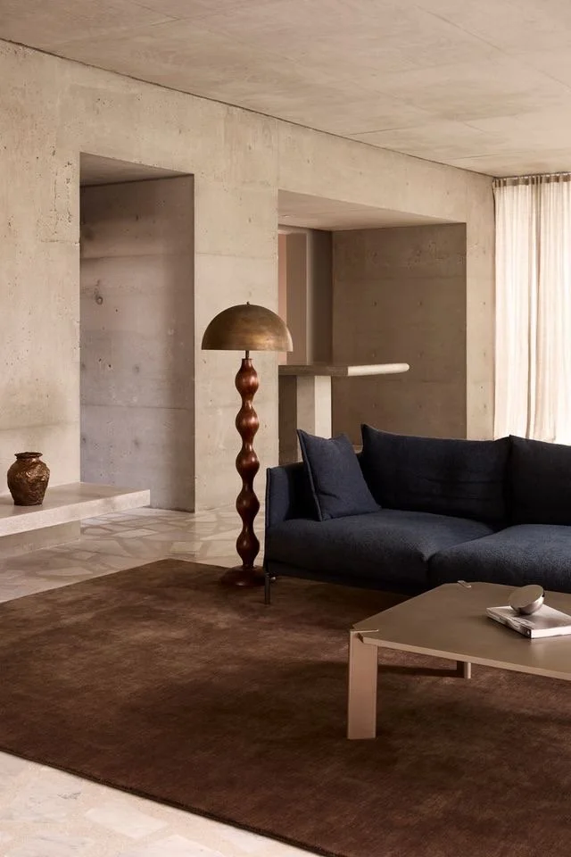Modern living room with concrete walls, a navy blue sectional sofa, a beige coffee table with a remote and magazine, a brown rug, a tall decorative floor lamp, and a neutral-colored window curtain.