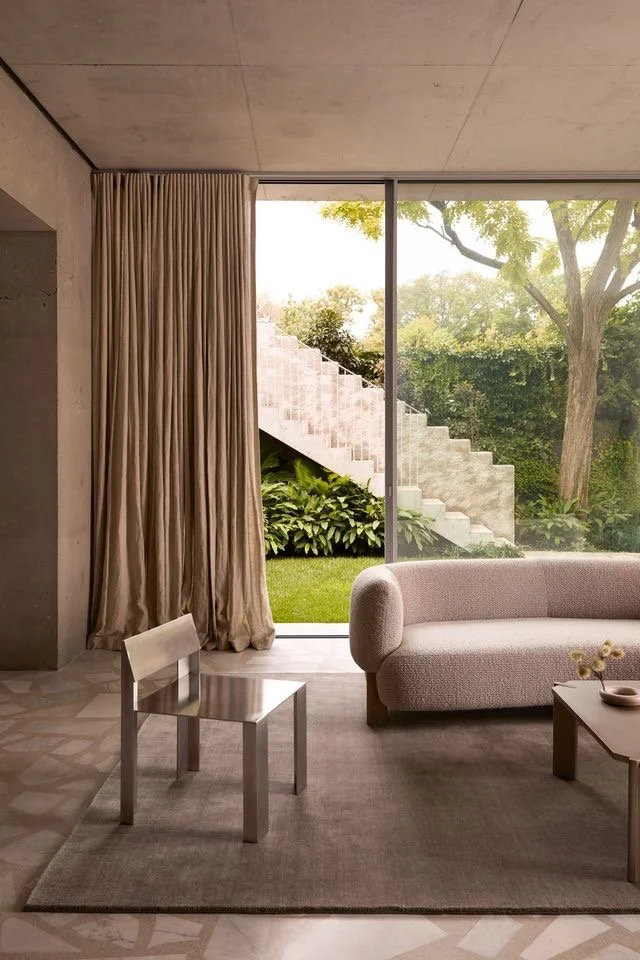 Modern living room with large glass window showing outdoor greenery and a staircase, beige curtains, a light pink sofa, a small chair, a coffee table with flower decoration, and a patterned rug.