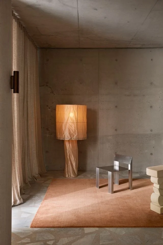 Minimalist room with concrete walls, a peach-colored rug on a marble floor, a tall stone table lamp, a small metallic chair, and beige-outlined curtains.