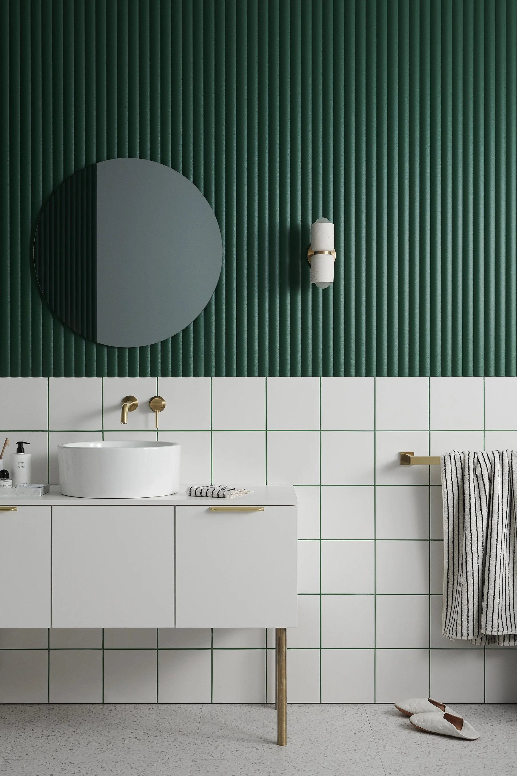 Modern bathroom with green tiled wall, white vanity with round vessel sink, round mirror, brass fixtures, white towel with black stripes, and white slippers.
