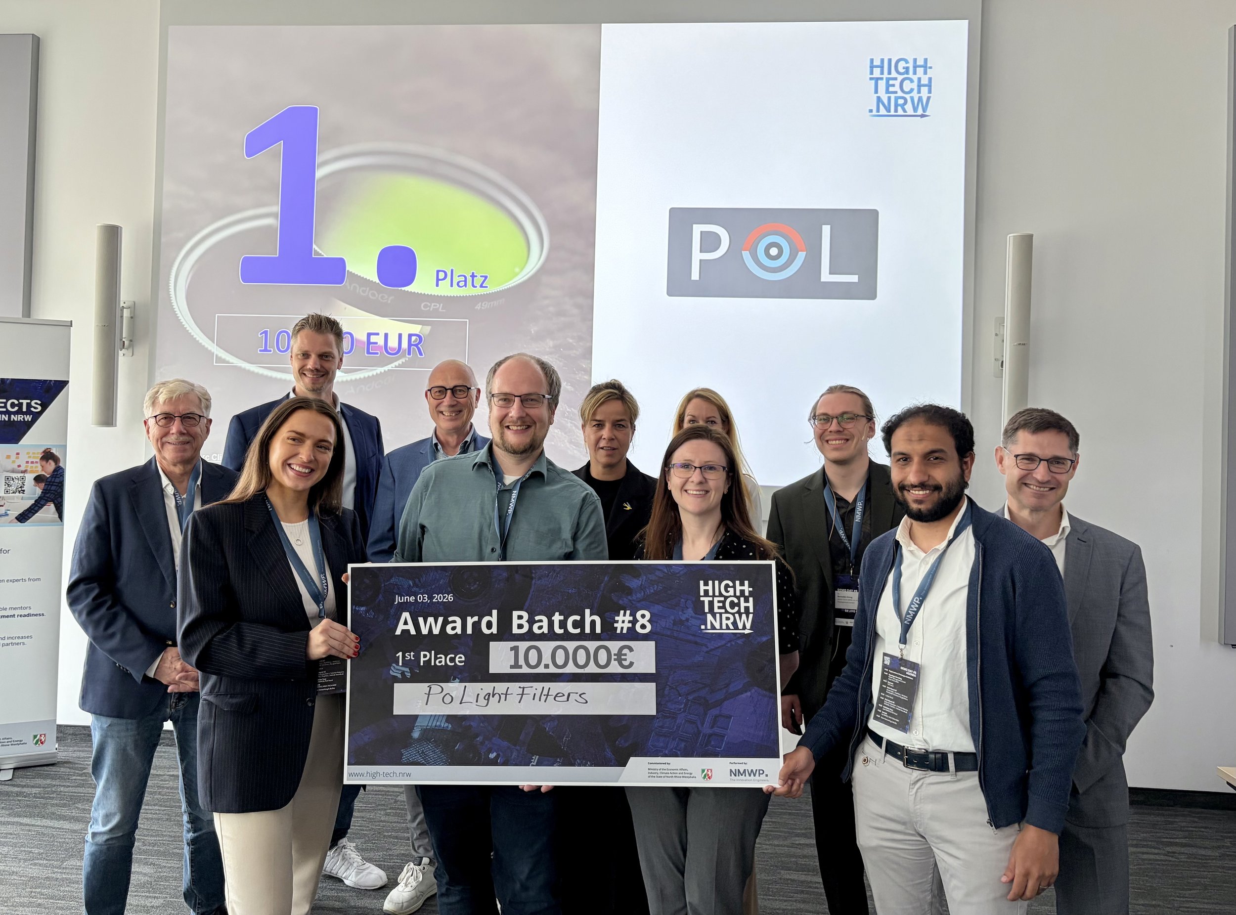 Group of people at an award ceremony holding a large check for 10,000 euros for first place, with a banner and presentation slide in the background indicating a high-tech event in NRW.