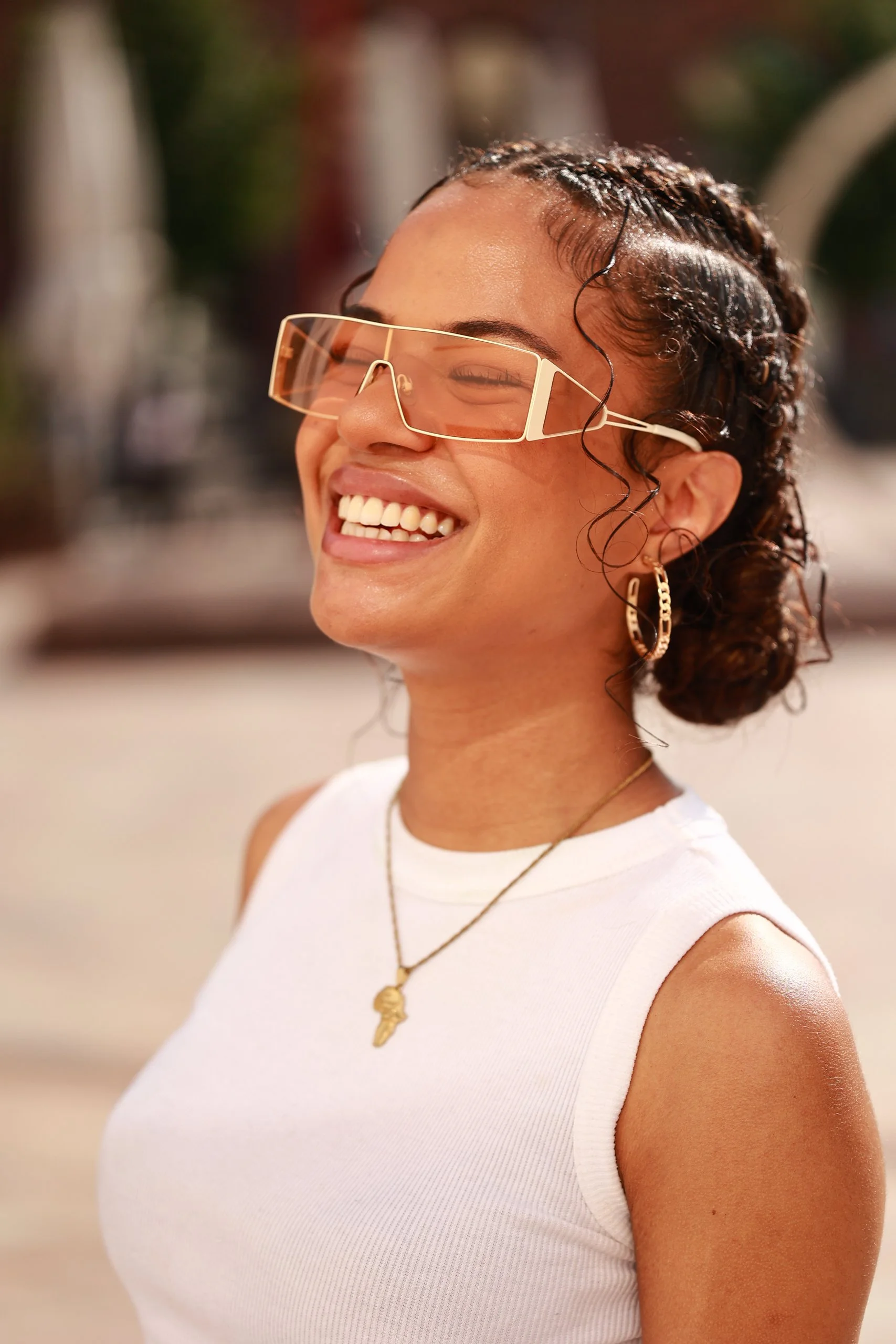 A woman smiling outdoors, wearing large orange-tinted sunglasses, gold hoop earrings, a gold necklace with a key pendant, and a white sleeveless top.