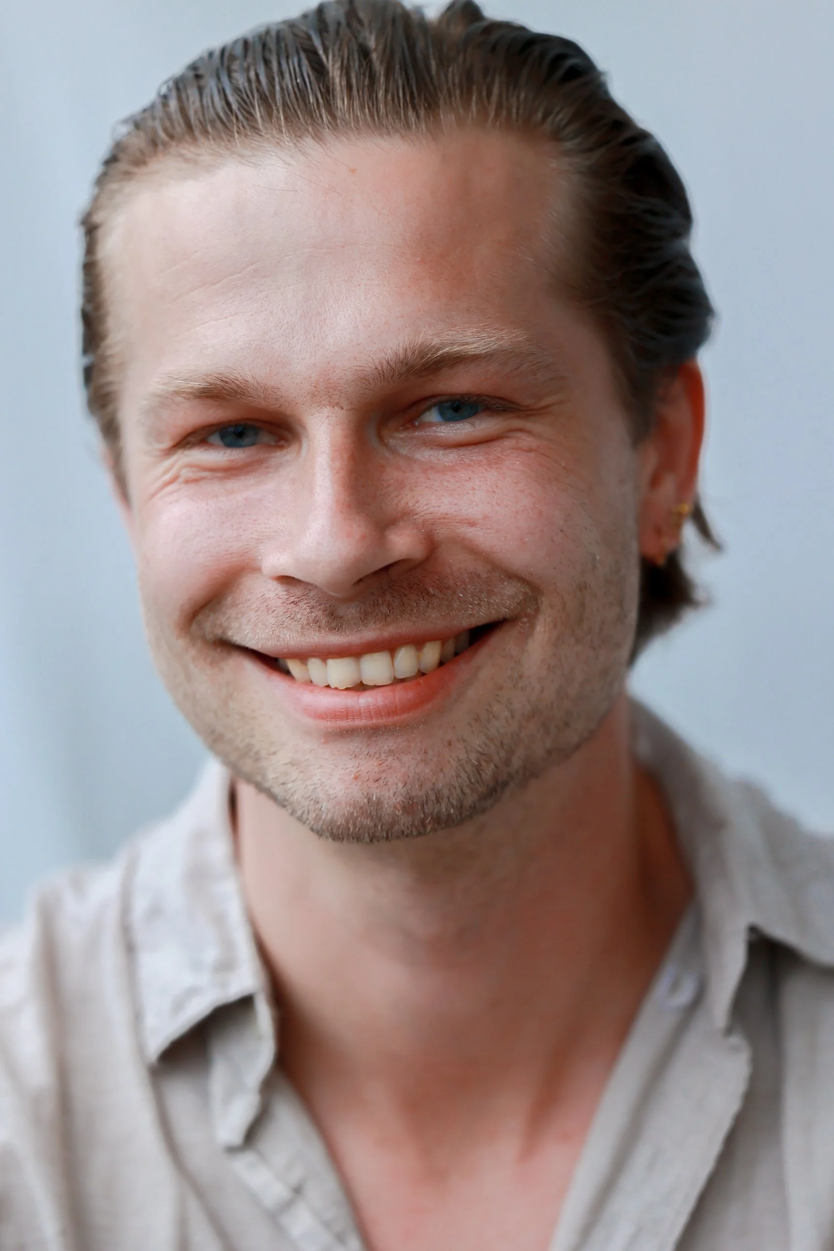 Close-up of a smiling man with blue eyes, short brown hair, and light facial hair, wearing a light-colored shirt.