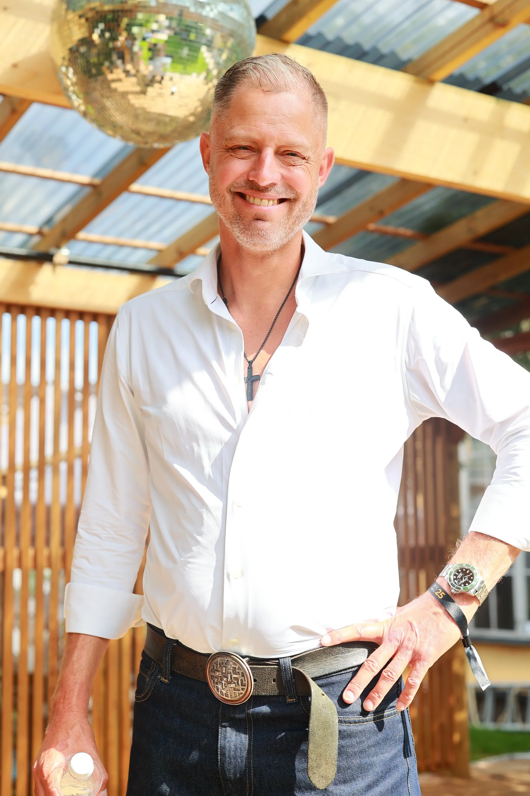 A smiling man with a white shirt, dark jeans, and a belt with a decorative buckle, standing outdoors under a roof with a disco ball hanging above him. He has short hair, a beard, and is wearing a watch and a cross necklace.