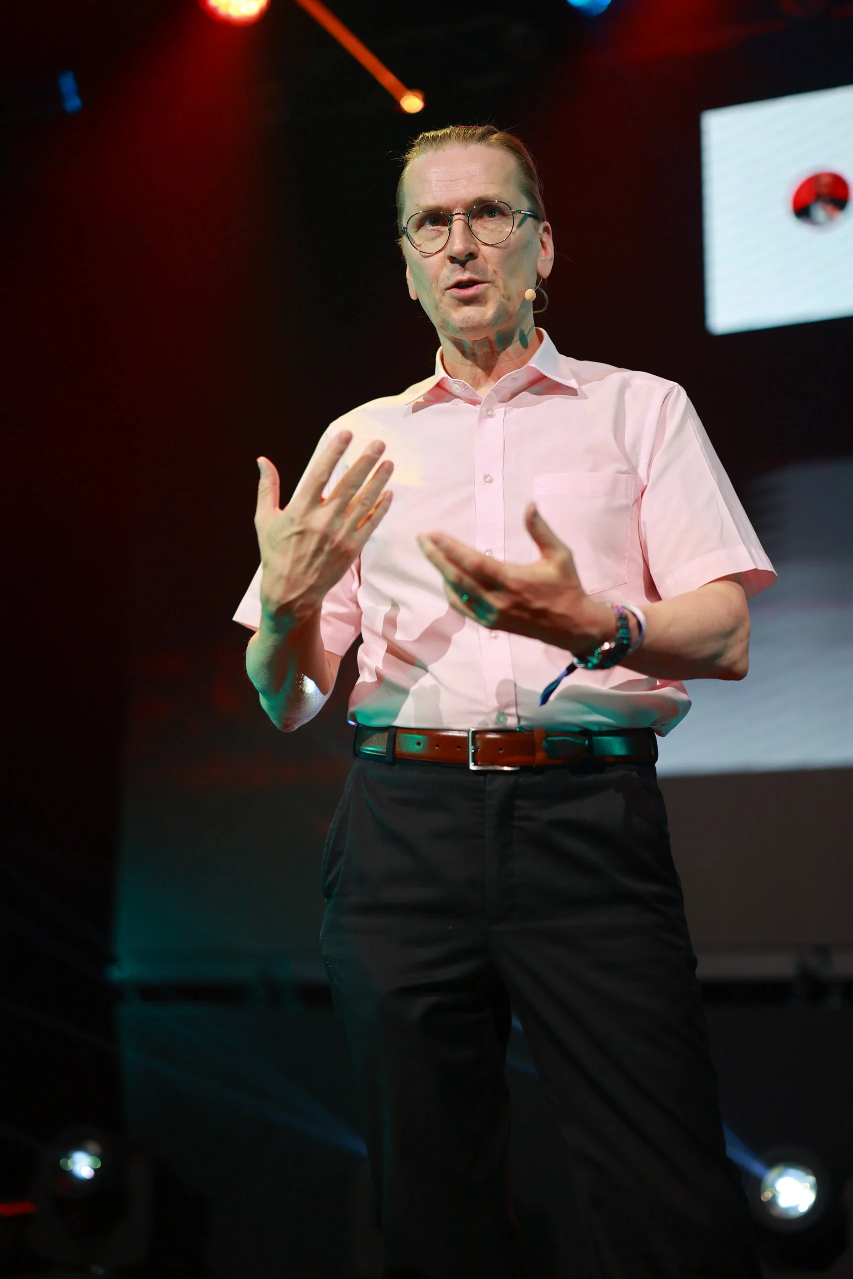 A man in a pink collared shirt and black pants, wearing glasses and a watch, speaking on a stage with a microphone headset, gesturing with his hands.