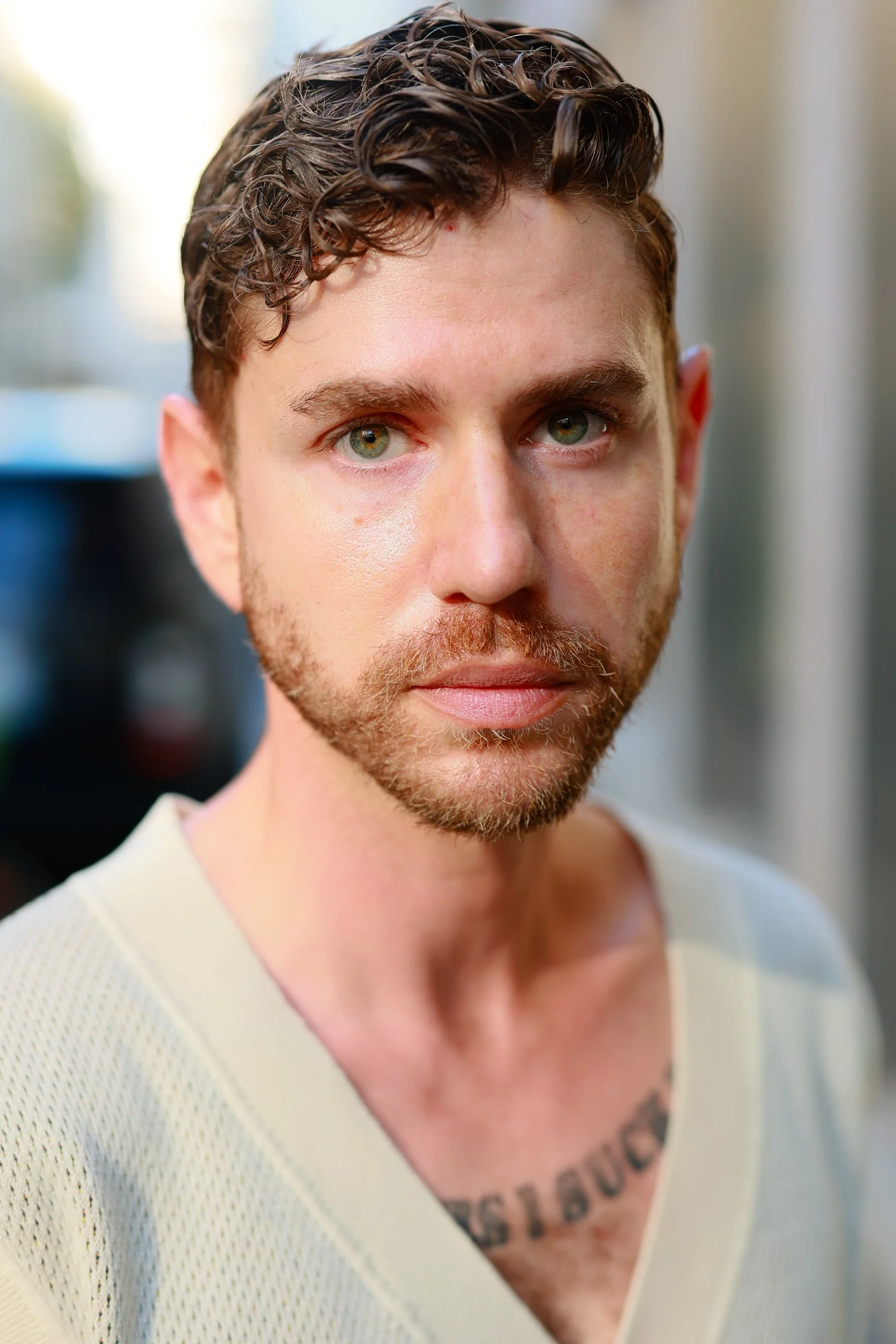 Close-up portrait of a young man with wavy brown hair, green eyes, and a beard, wearing a light-colored shirt.