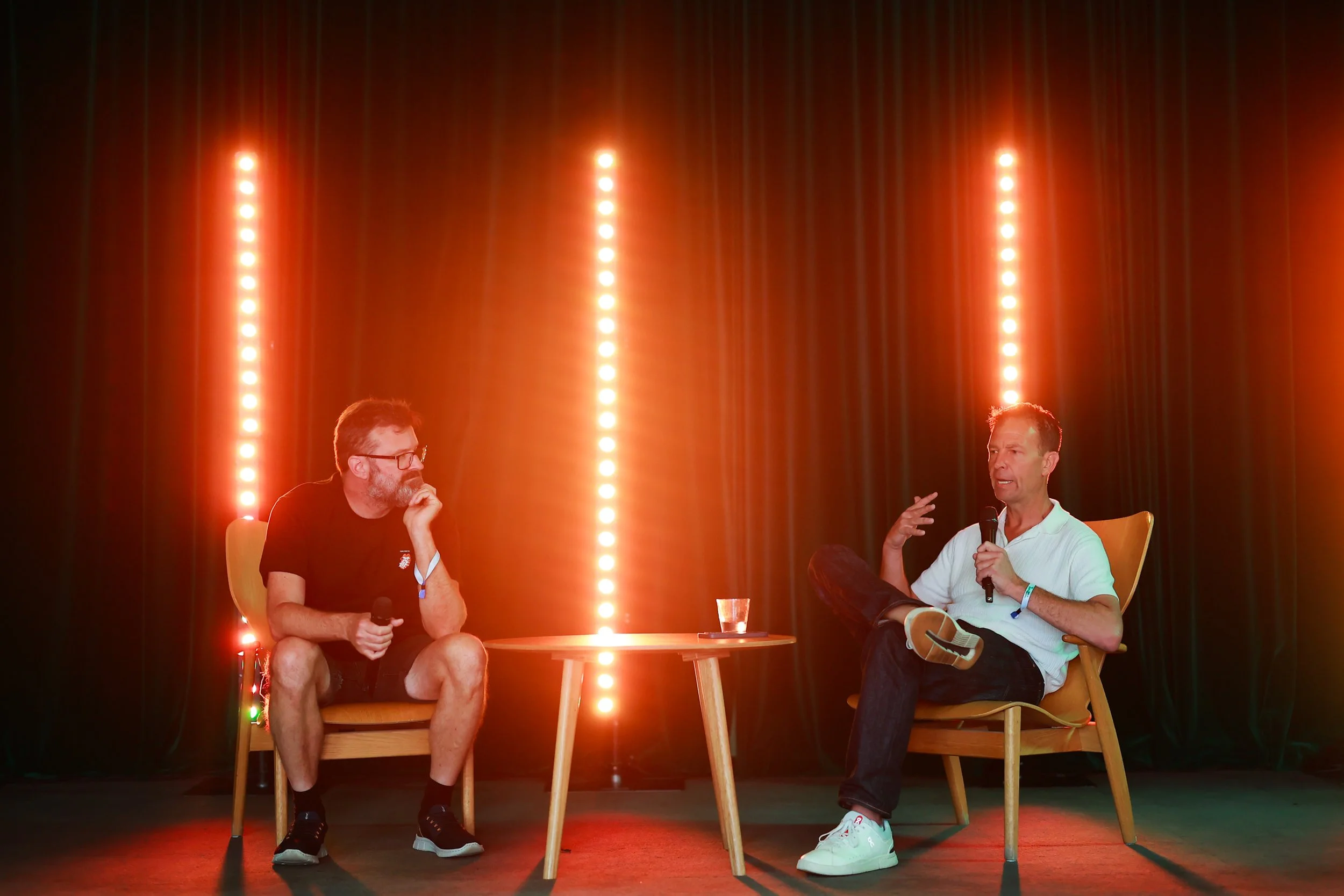 Two men sitting on stage engaging in a conversation with microphones, black curtains, and vertical red stage lights behind them.