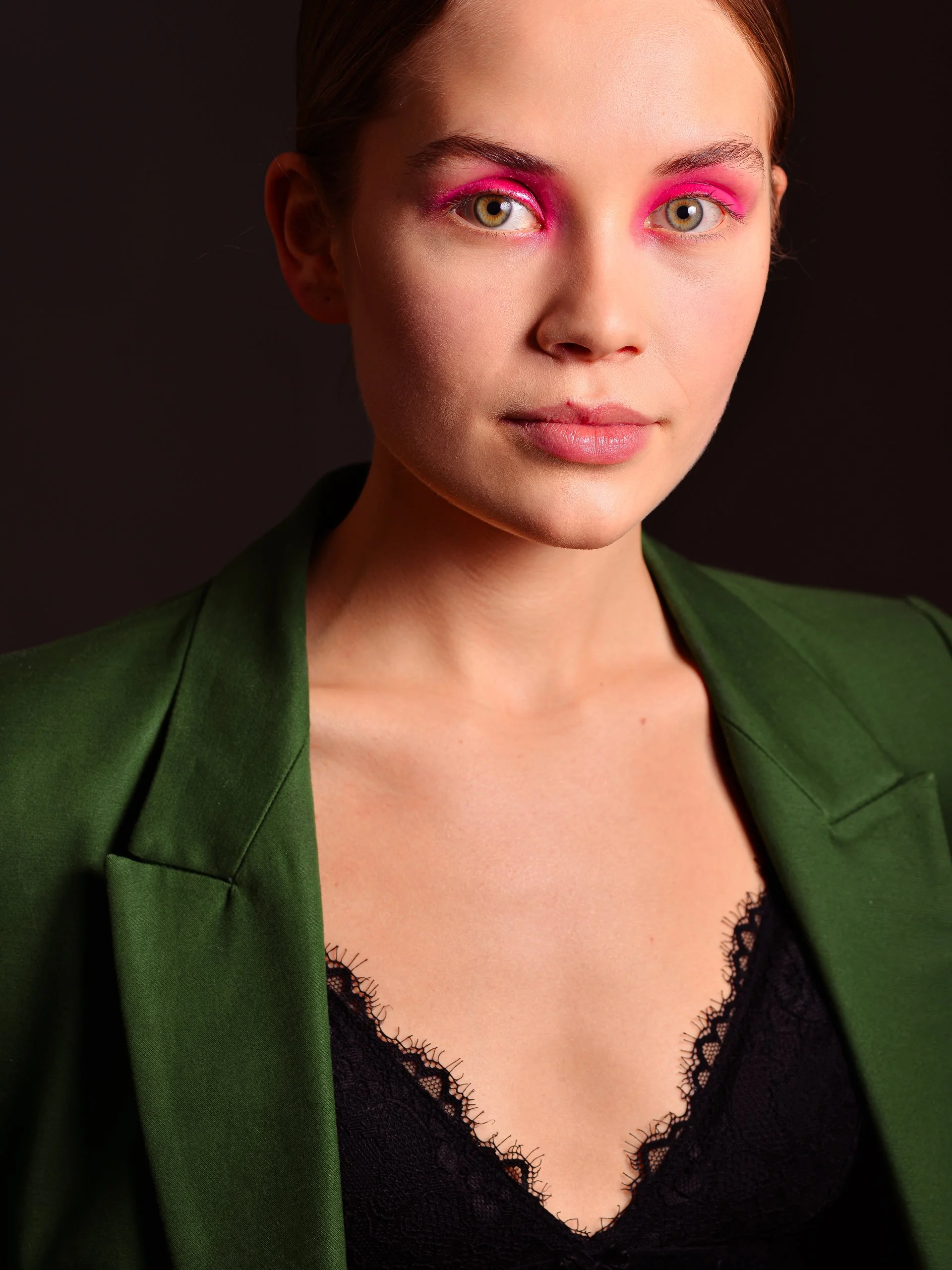 A woman with light skin and blonde hair with pink eyeshadow, wearing a green blazer over a black lace top, against a dark background.