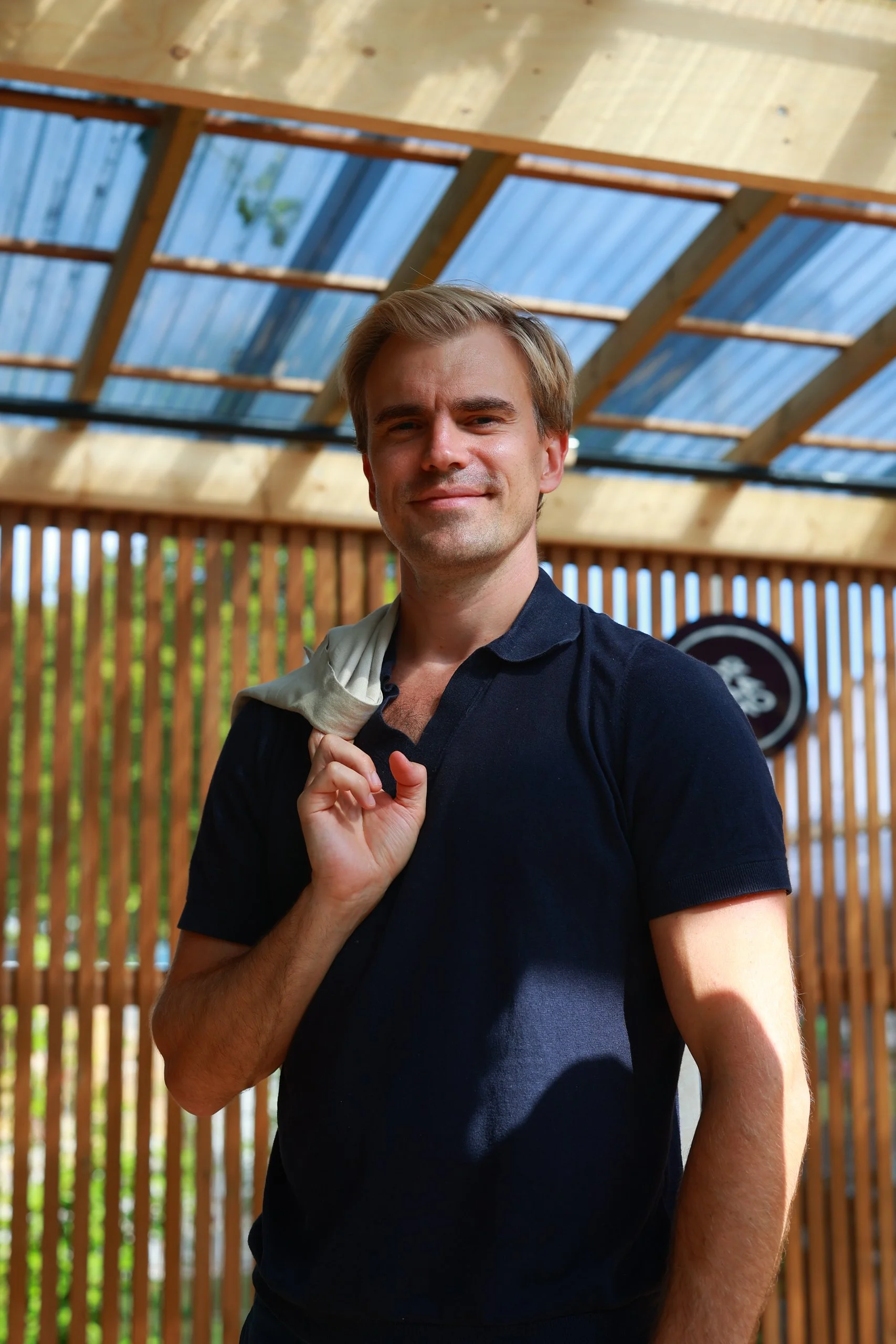 A man with blonde hair holding a cloth draped over his shoulder, standing inside a wooden structure with a skylight ceiling, smiling at the camera.