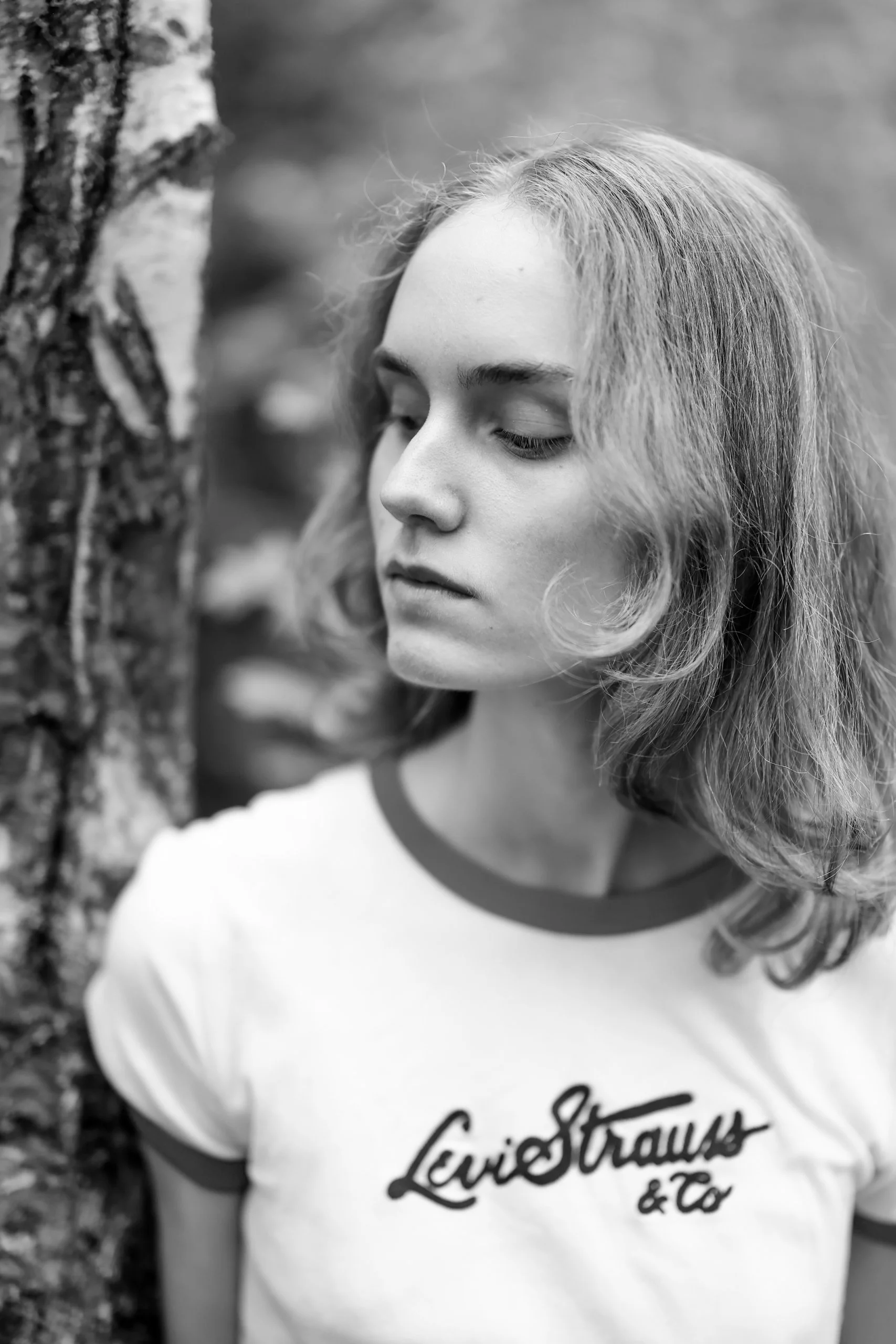 Black and white photo of a young woman with medium-length wavy hair wearing a white T-shirt with ringer collar and cursive text, standing next to a tree.