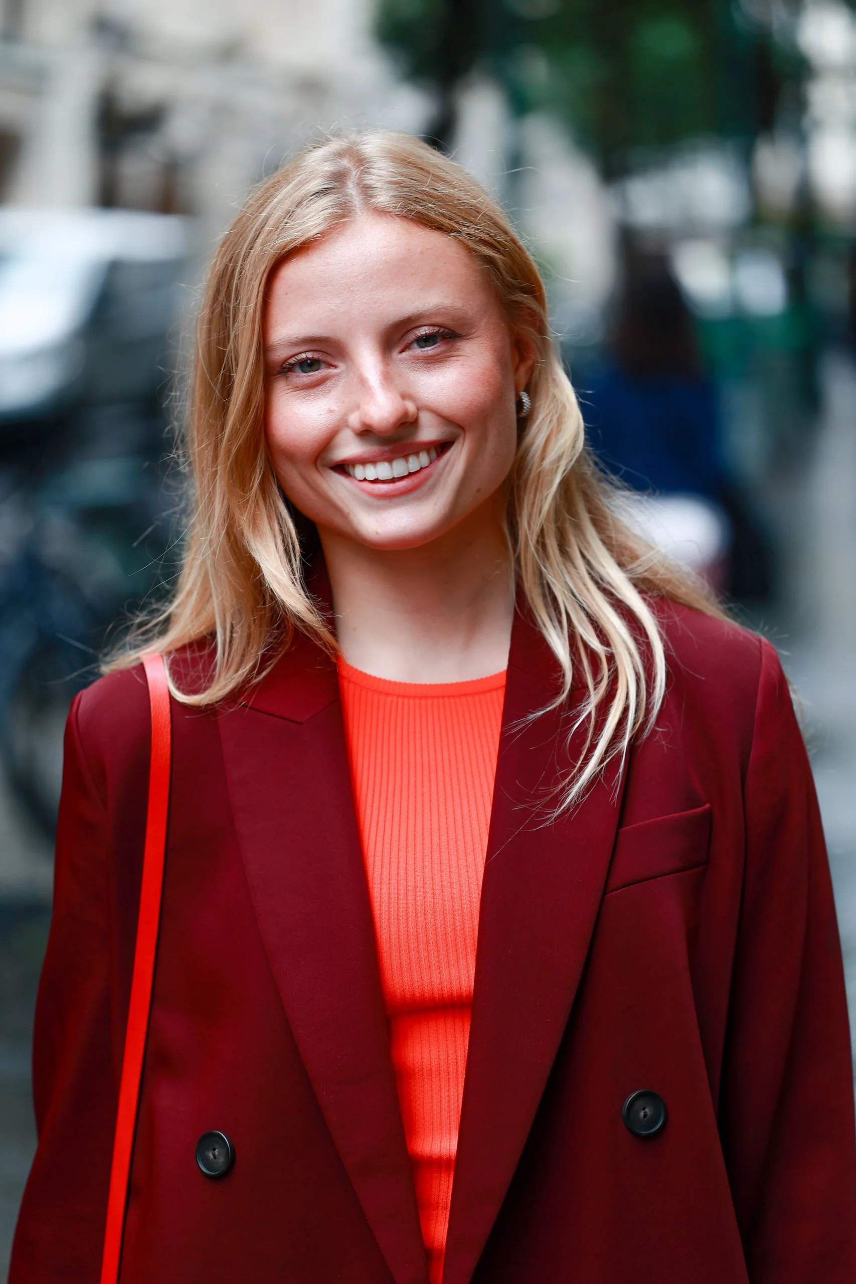 A smiling woman with blonde hair wearing a red blazer and a red top, standing outdoors in a city street setting.