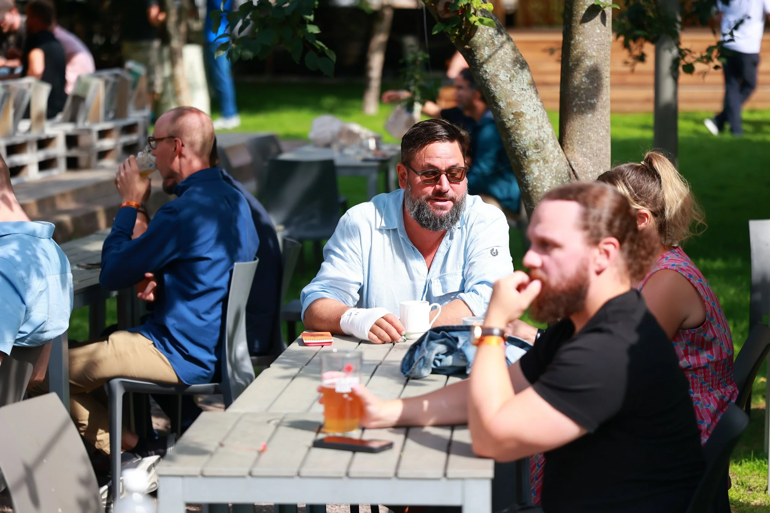 People sitting and socializing outdoors at a garden party or restaurant, some holding drinks and coffee cups, with trees and greenery in the background.