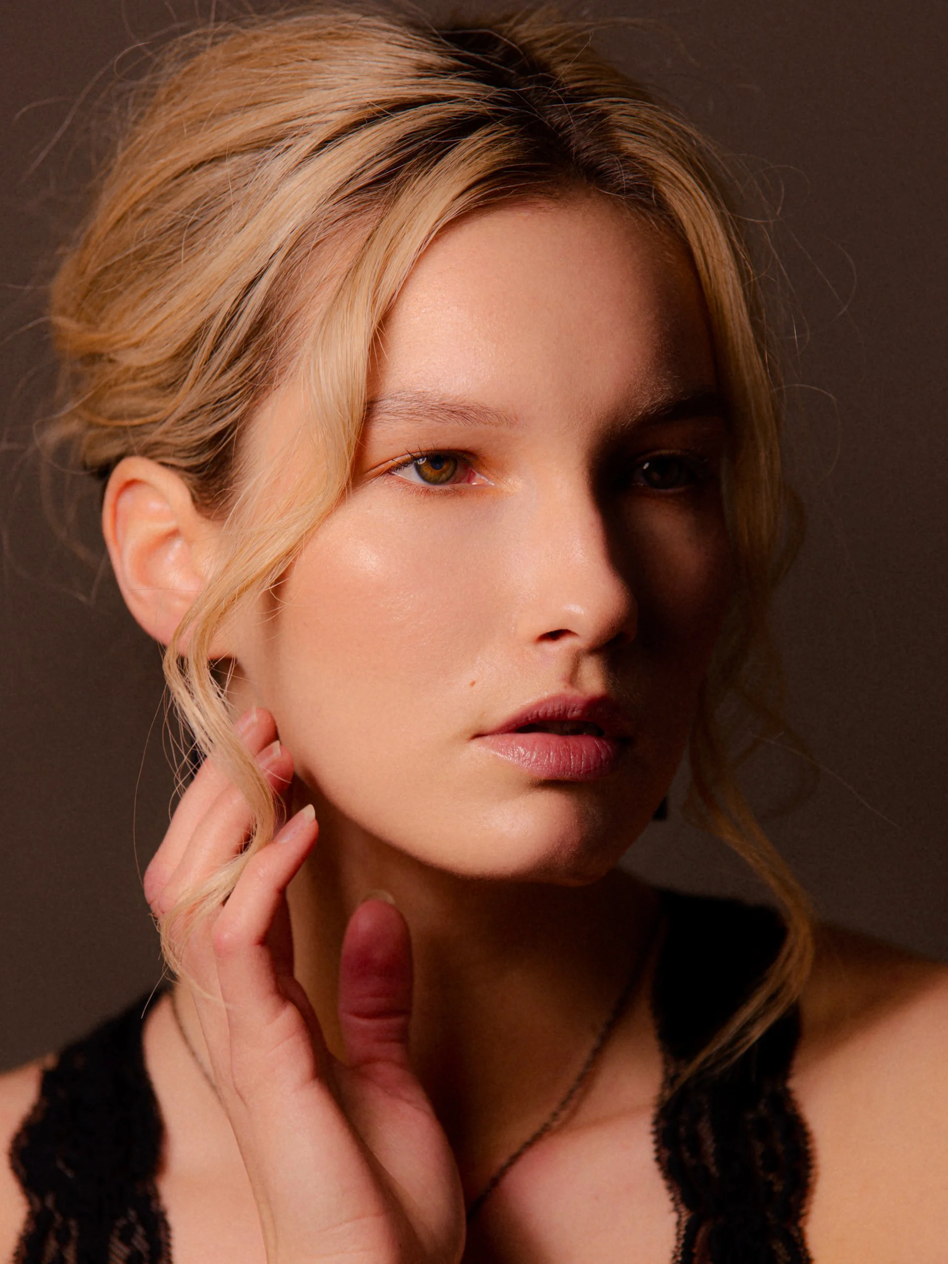 Close-up portrait of a young woman with blonde hair, posing with her hand near her cheek, wearing black lace clothing and a necklace.