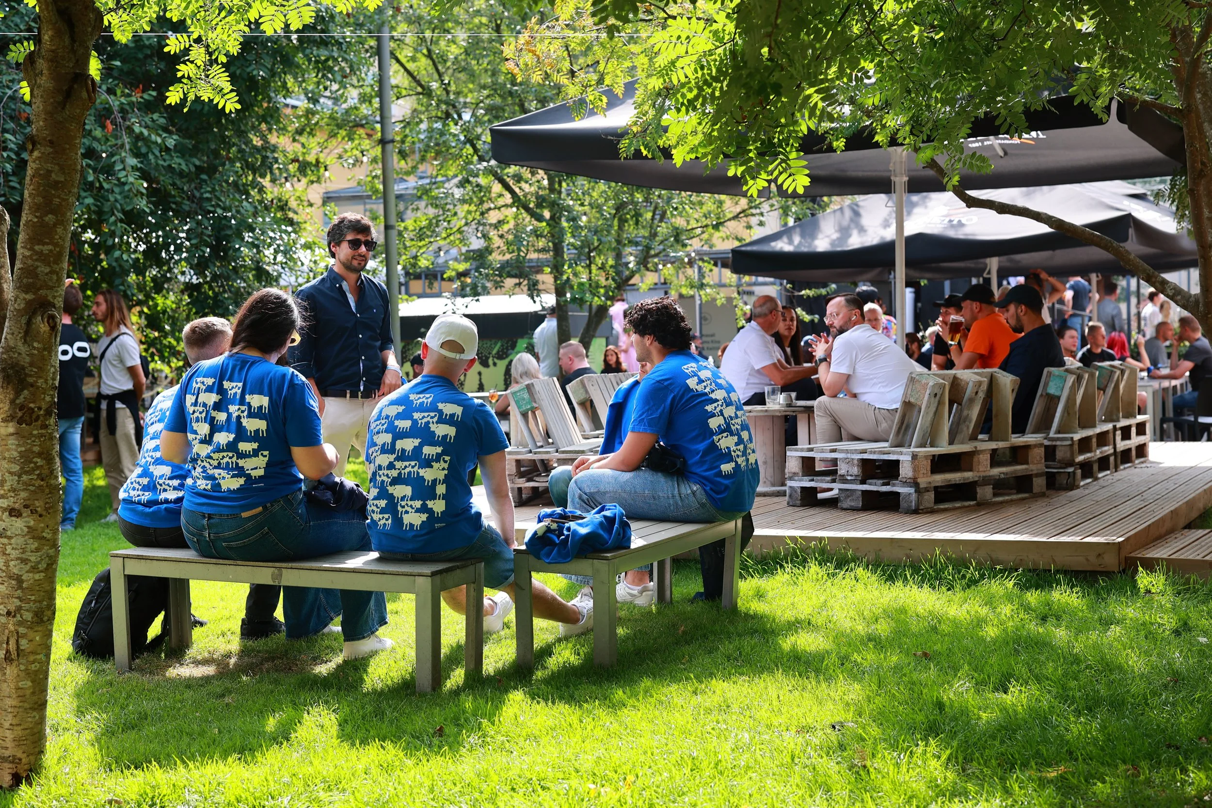 People gather outdoors at a park or patio with trees, benches, and umbrellas, socializing and enjoying a sunny day.