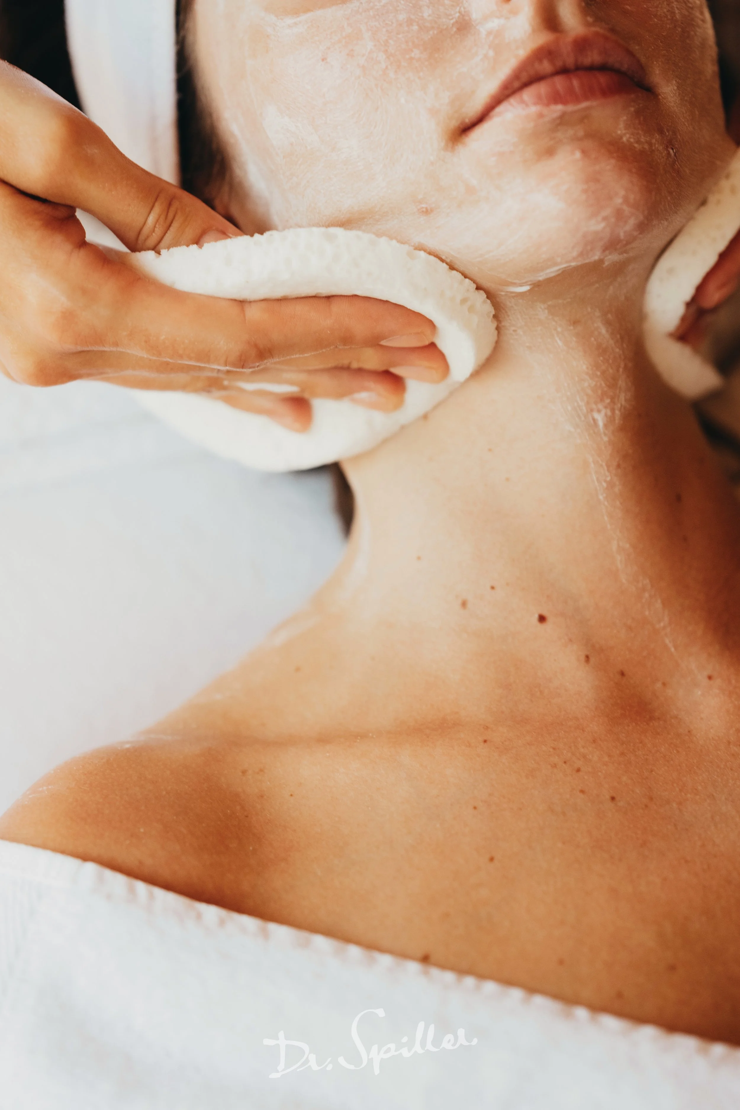 A person receiving a facial treatment, with a skincare specialist using a white sponge on their face and neck.